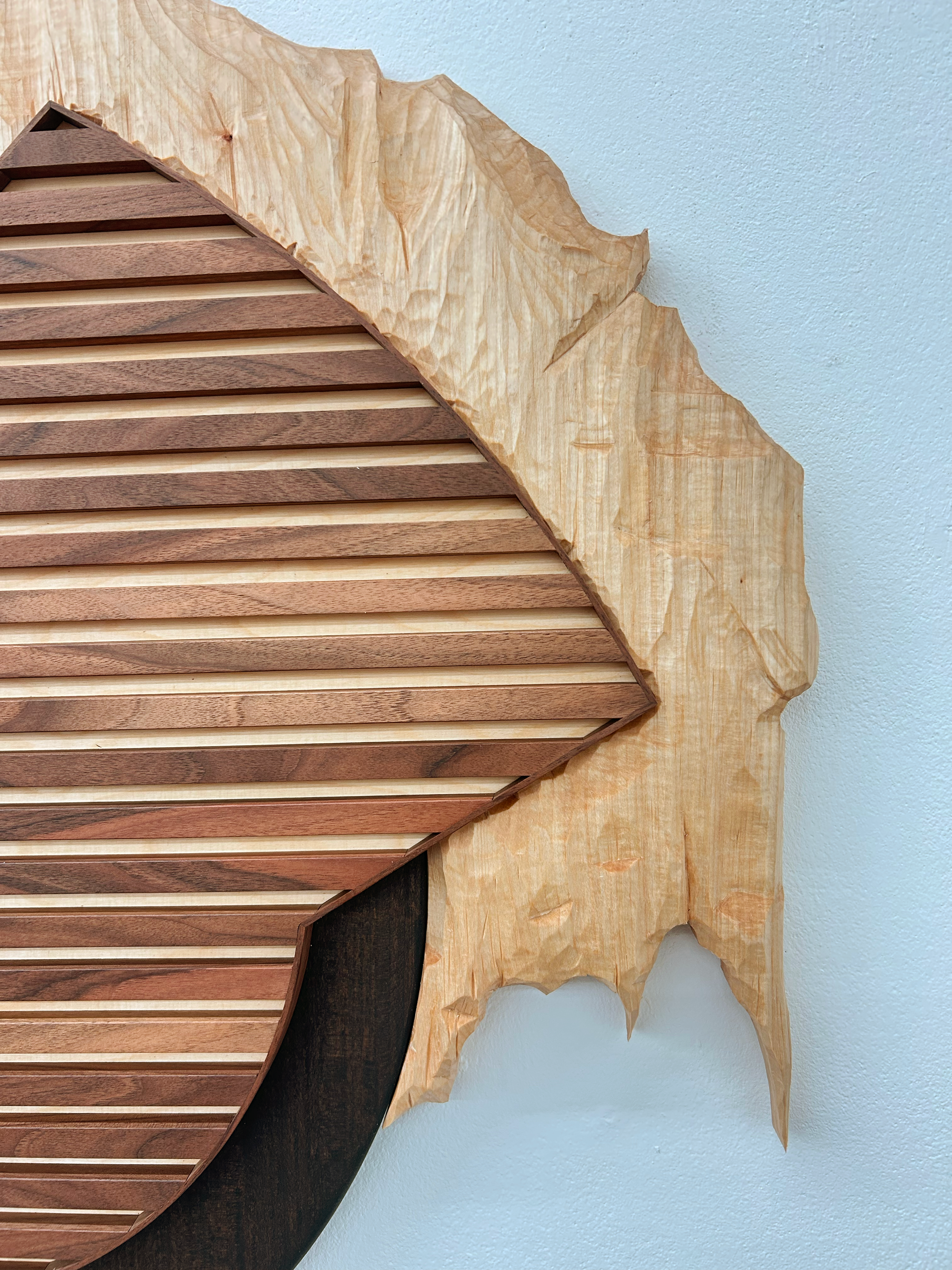 Von Julian Schomäker gearbeitete Skulptur als Wandobjekt. Komposition verschiedener Holzelemente. Es ist ein geschnitzter Berg, in den ein Lamellenartiges Objekt eingelassen ist, das auf einem schwarzen Halbkreis sitzt, der auch an den Berg angrenzt.