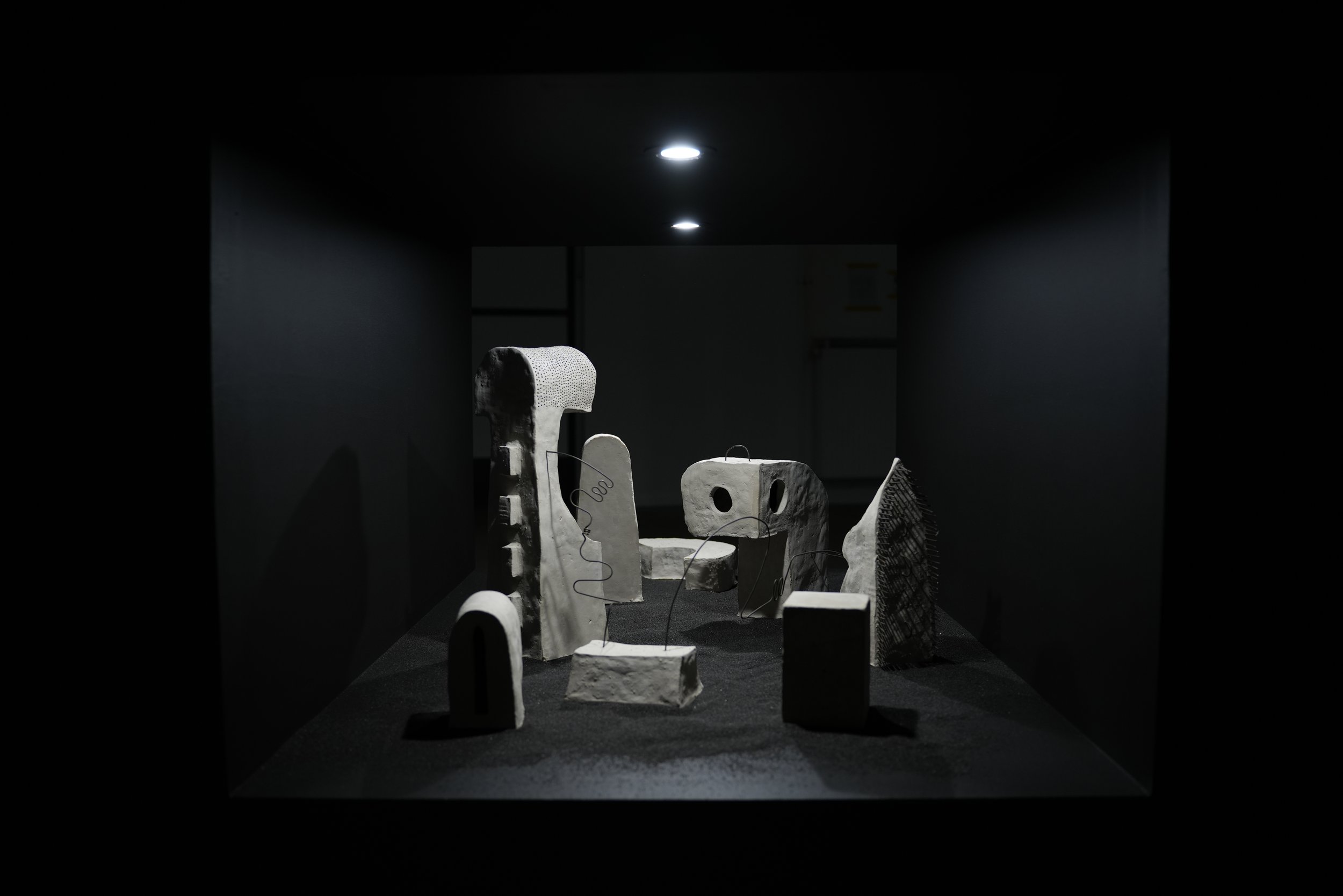 Landschaftliche Installation verschiedener, durch Julian Schomäker gefertigte, roh belassener Tonskulpturen. Die Skulpturen sind durch Drähte miteinander verbunden und sind auf schwarzem Sand in dem Durchbruch einer Wand installiert.