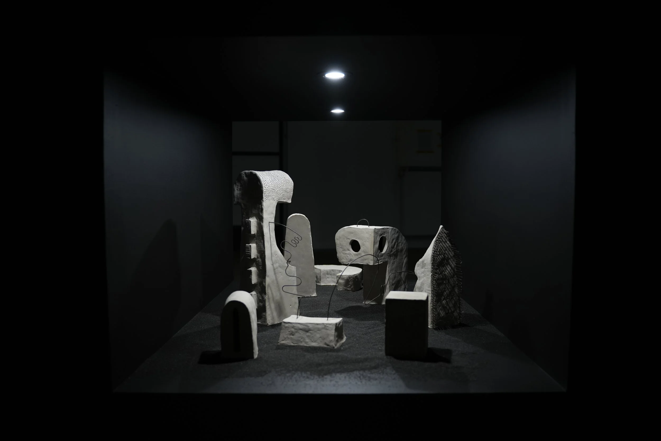 Landschaftliche Installation verschiedener, durch Julian Schomäker gefertigte, roh belassener Tonskulpturen. Die Skulpturen sind durch Drähte miteinander verbunden und sind auf schwarzem Sand in dem Durchbruch einer Wand installiert.