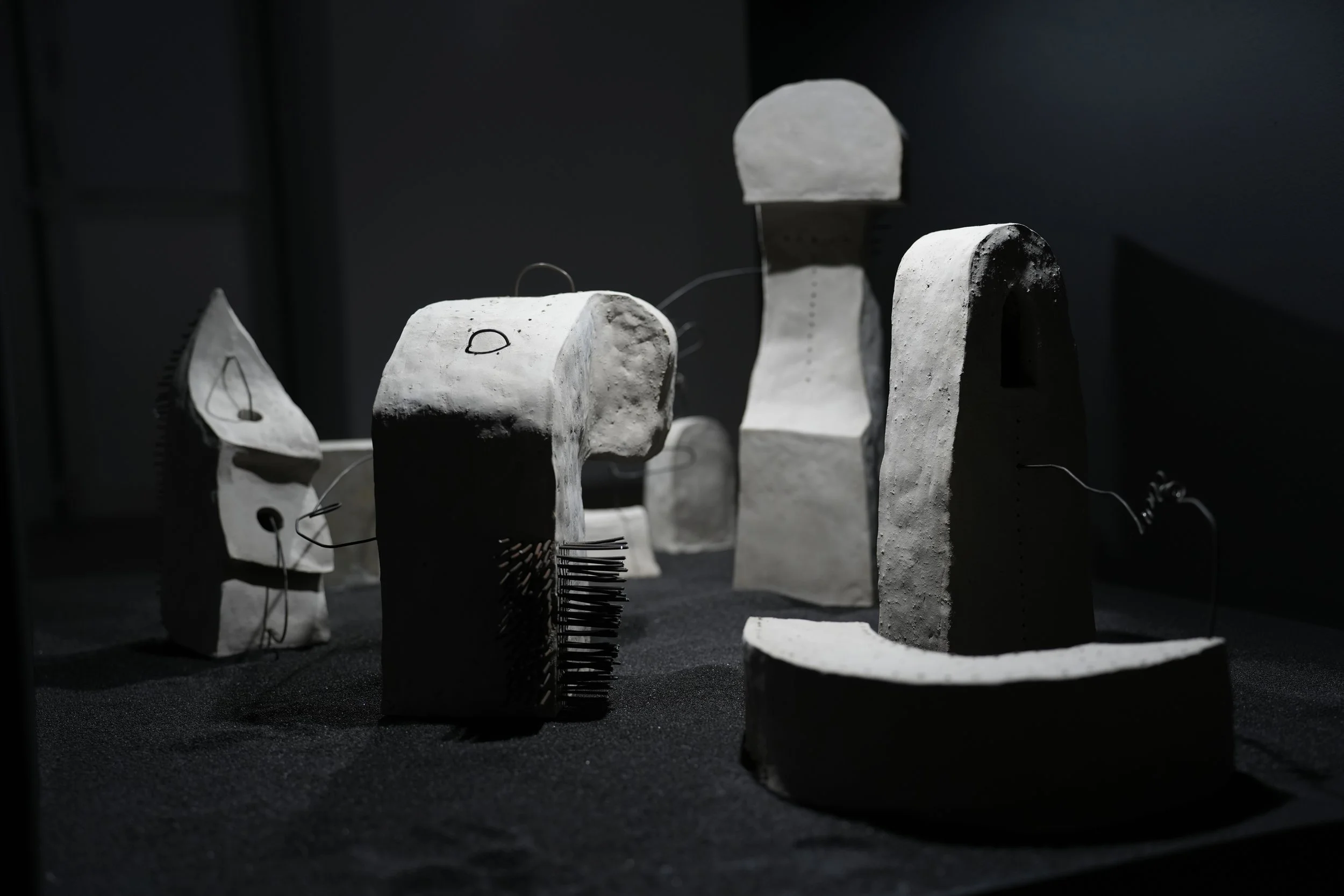 Zu sehen ist die landschaftliche Installation verschiedener, durch Julian Schomäker, gefertigter, roh belassener Tonskulpturen. Die Skulpturen sind durch Drähte miteinander verbunden und sind auf schwarzem Sand in einem Kasten installiert.