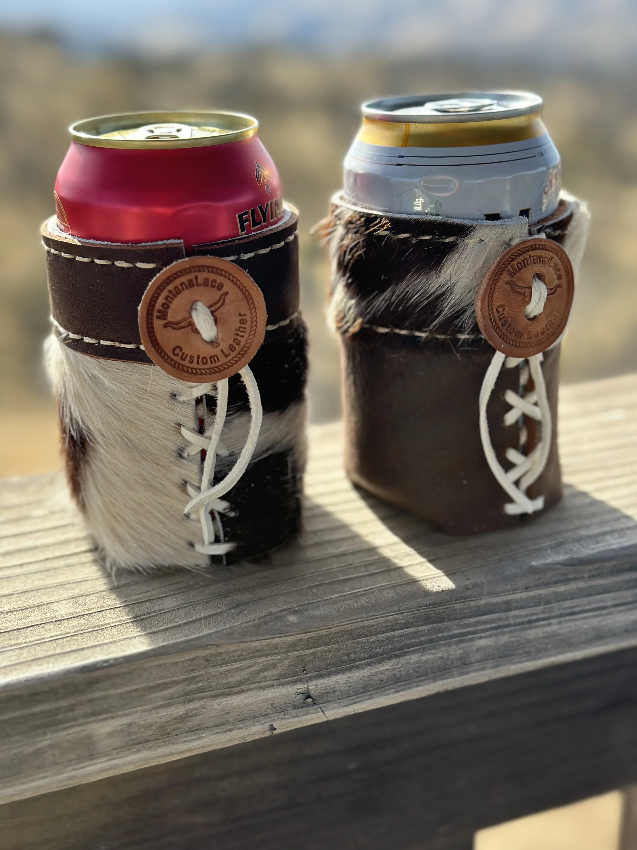 Two cans, one red and one white, are placed inside leather holders with cowhide accents and tags that read "MonaLace Custom Leather." They are resting on a wooden surface.
