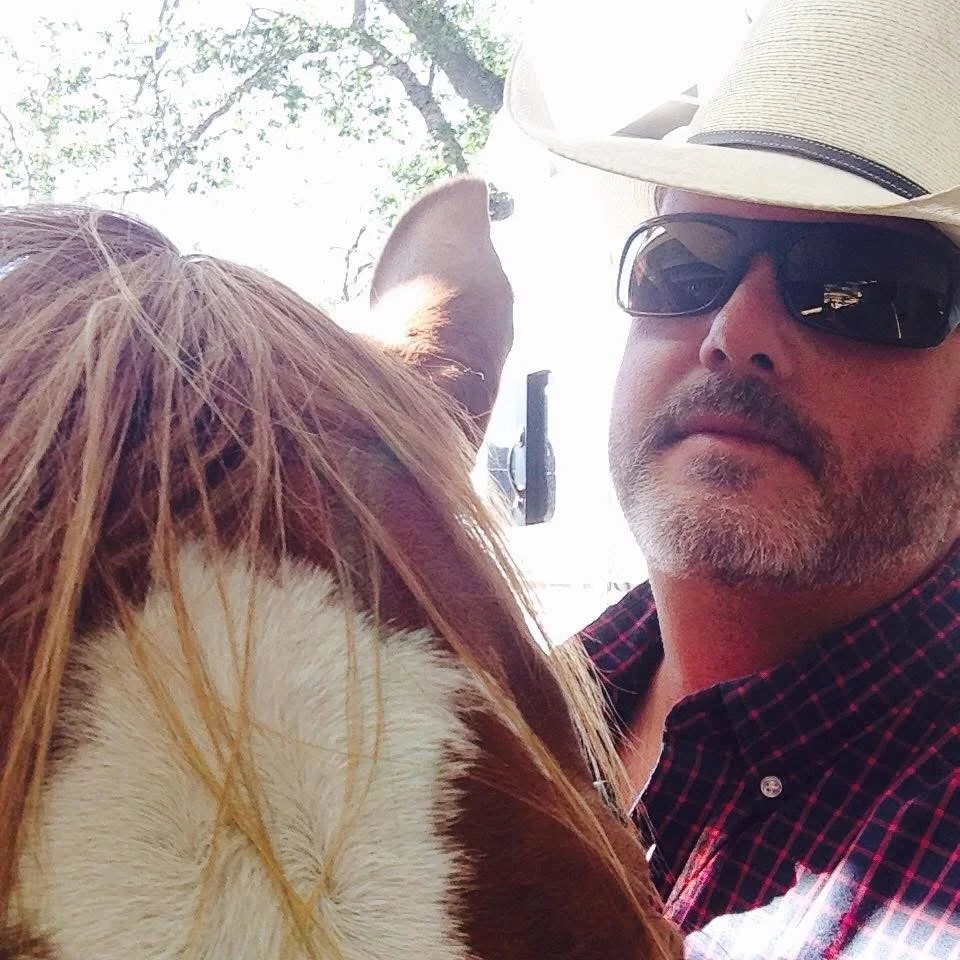 A man wearing sunglasses, a white hat, and a red plaid shirt takes a selfie with a horse. The horse has a brown and white coat and is close to the camera, partially blocking the view of the man.
