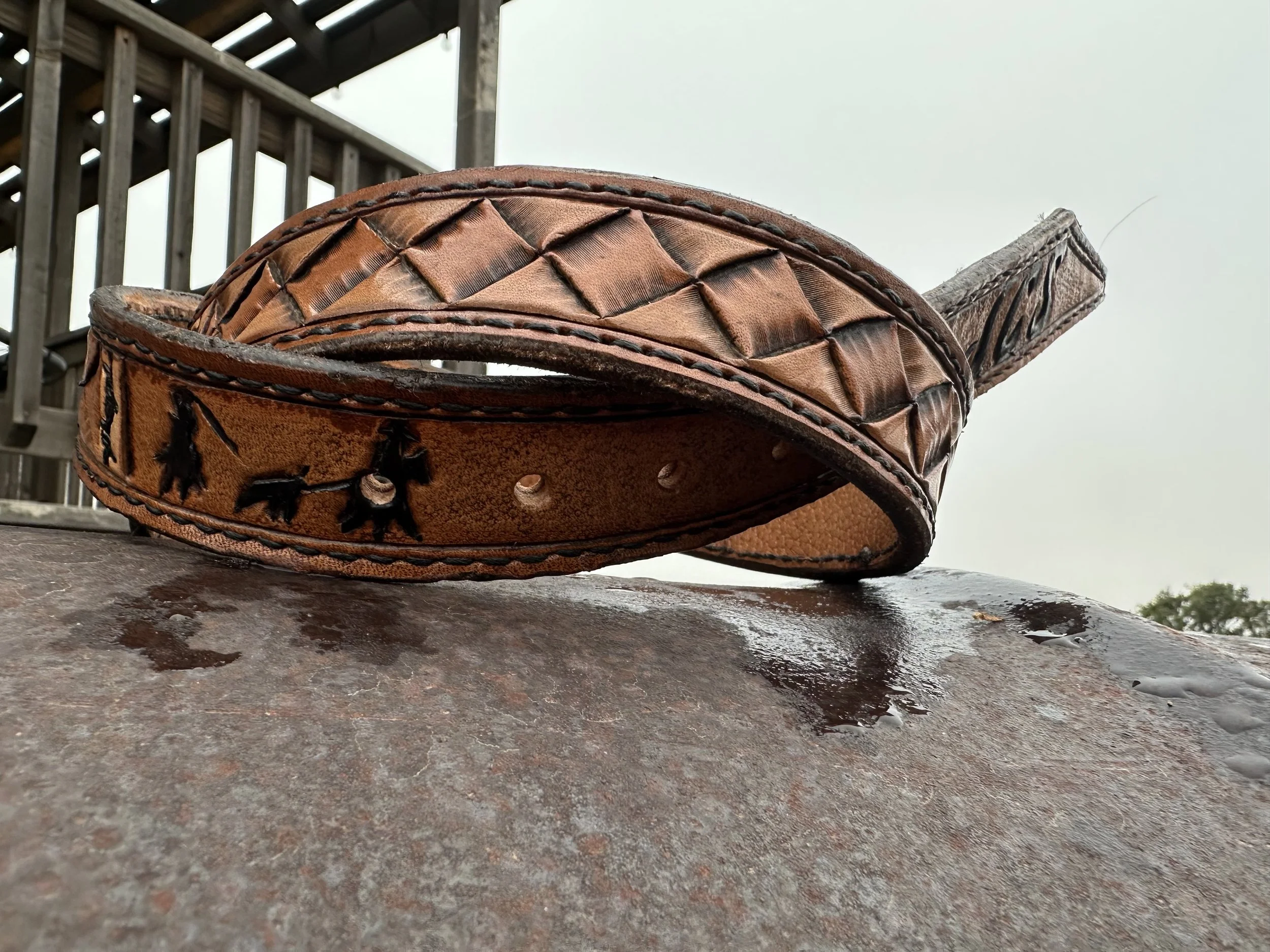 Close-up of a worn leather cowboy hat with decorative stitching, resting on a wet, rusty metal surface outdoors, with a background of a weathered wooden deck and cloudy sky.