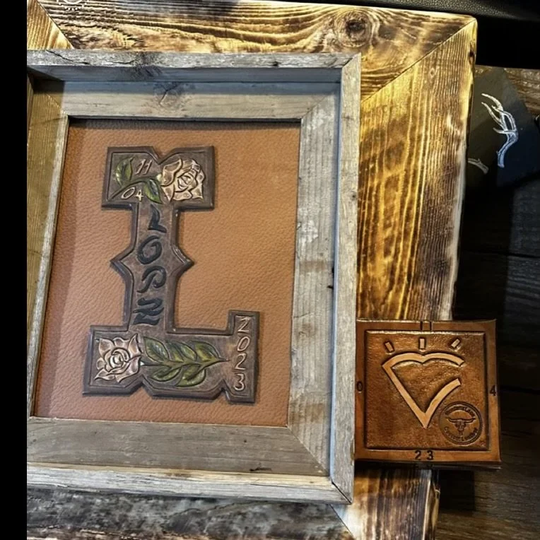 Decorative wooden crosses with intricate designs and engraved text. One cross has the words "In Loving Memory" and the years 1947-2023, and is placed inside a rustic wooden frame. The other small wooden piece features a heart with a smaller circle in