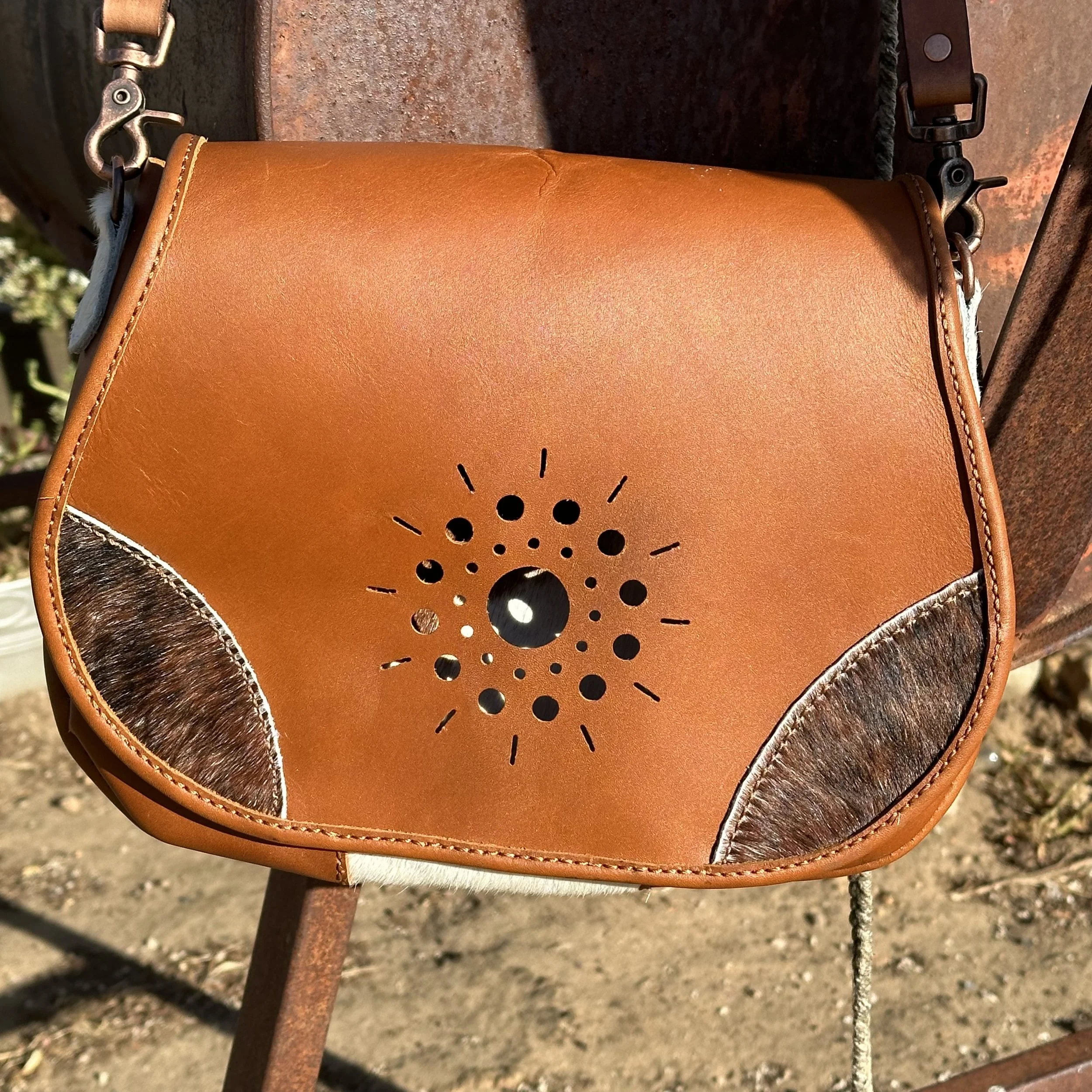 Close-up of a tan leather saddle pad with black and brown trim, attached to a saddle with metal hardware and a rope on a rusty metal stand outdoors.