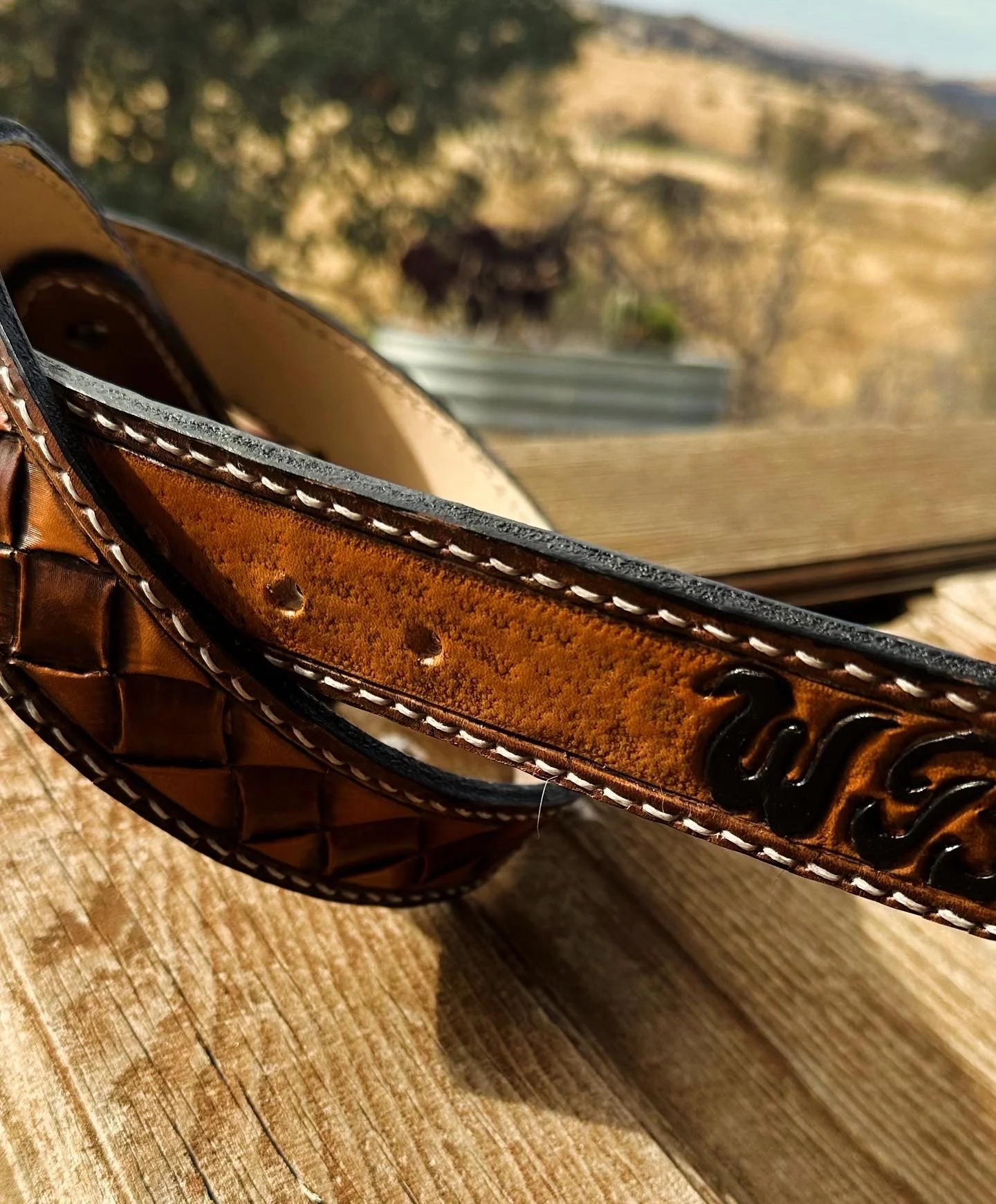 Close-up of a brown leather belt with white stitching and branding, resting on a wooden surface with a rustic background.