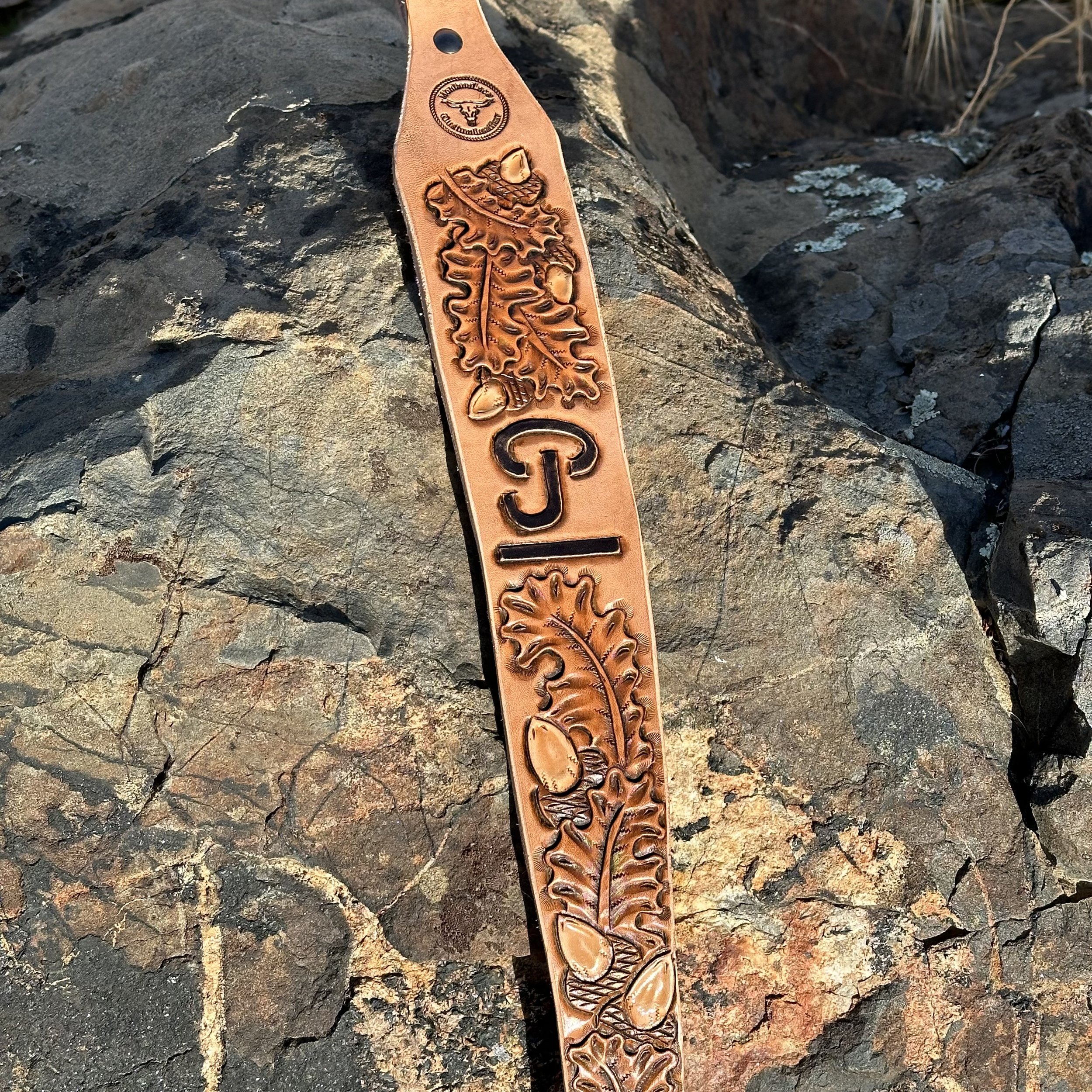 Leather strap with engraved designs including a snake and a deer's head, placed on a dark rocky surface.