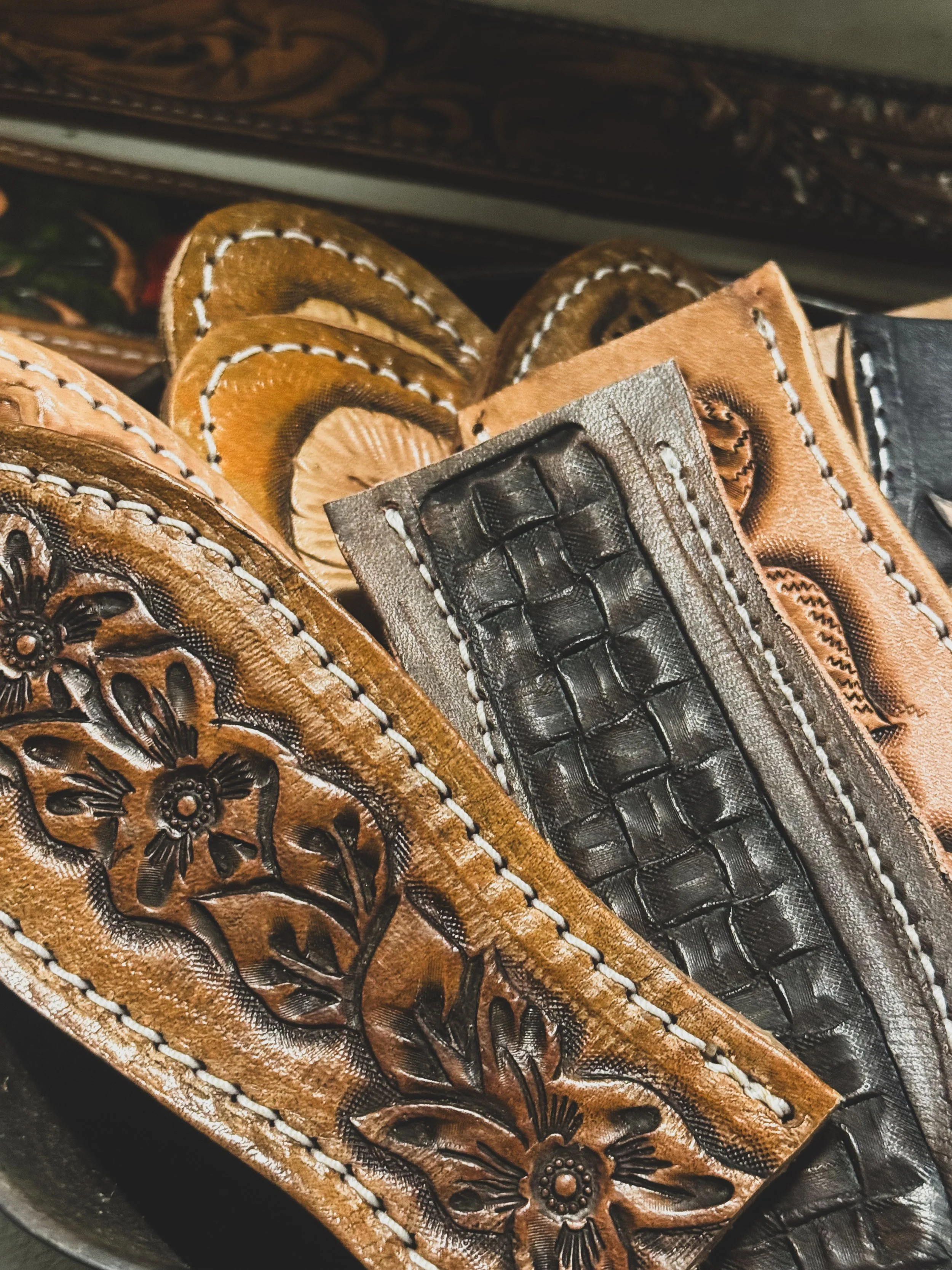 Close-up of a stack of leather cast iron pot holders in various colors, including brown, black, and tan, with embossed floral patterns and white stitching along the edges.