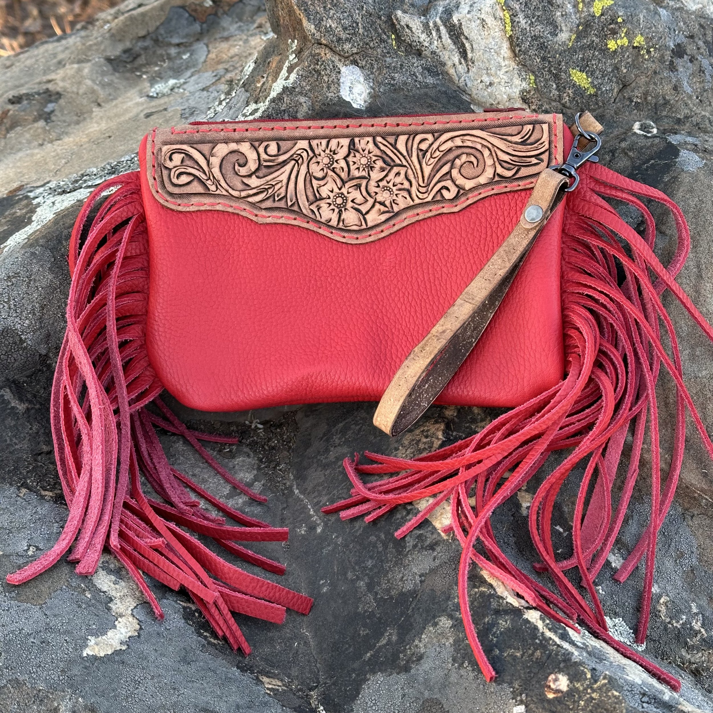 Close-up of a red fringed handbag with a decorative leather flap engraved with floral and swirling designs, resting on a rock surface.