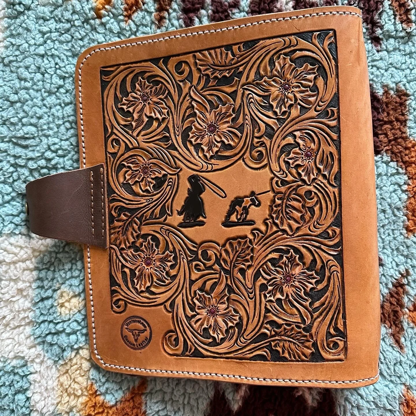 Leather folder with intricate floral and animal cutouts, featuring a steer head logo at the bottom left, lying on a multicolored textured rug.