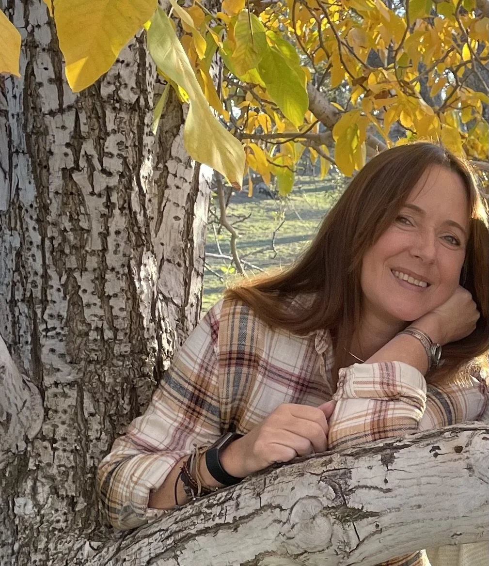 A woman with brown hair lying on a tree branch, smiling, surrounded by autumn yellow leaves.