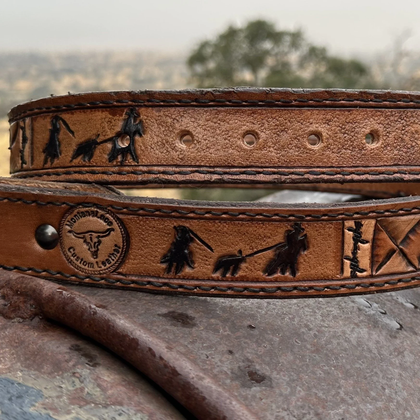 Close-up of a brown leather belt with embossed horse and rider silhouettes, resting on a rusted metal surface with a blurry outdoor background.