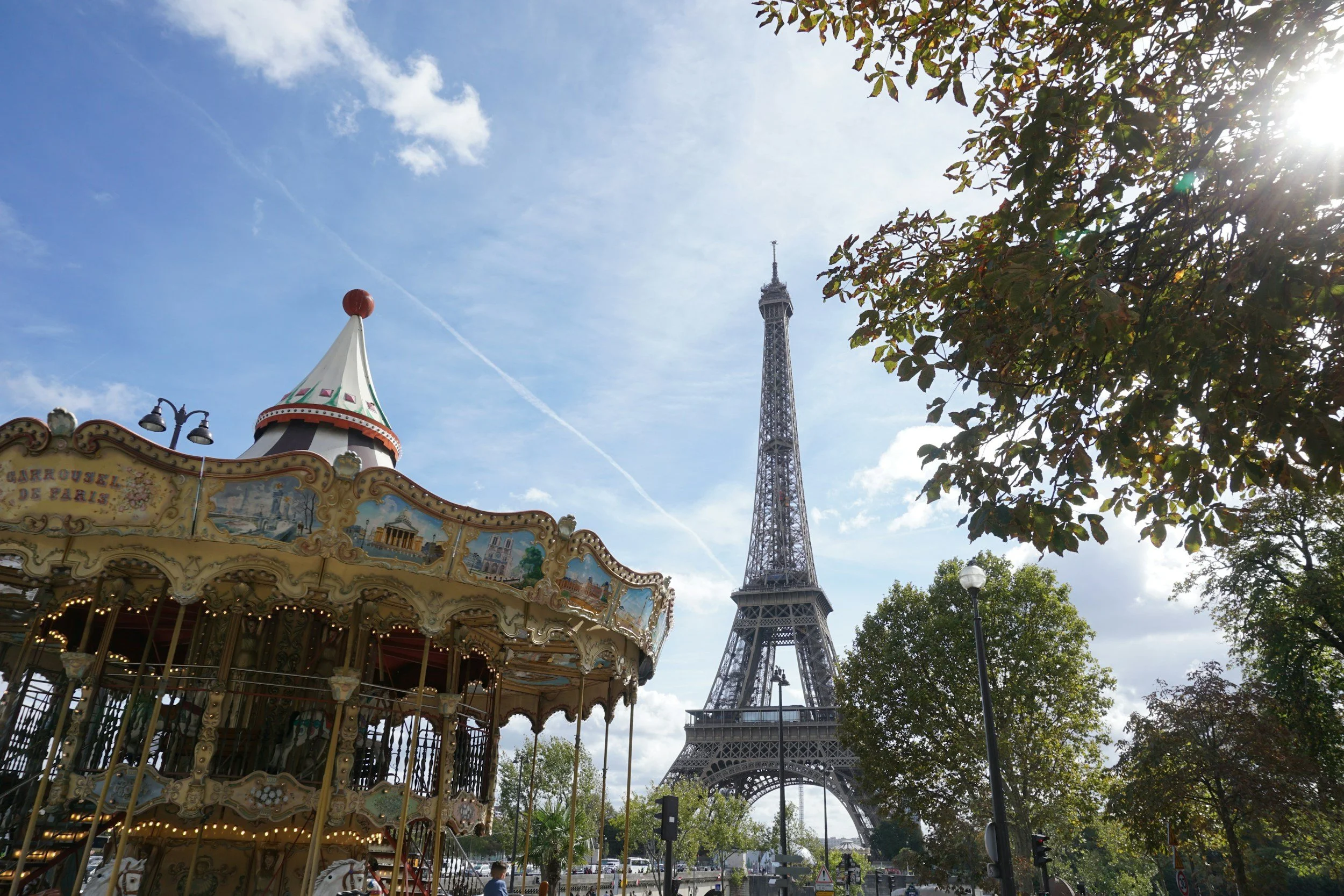 A carousel ride is in the foreground with decorative murals, and the Eiffel Tower is in the background with partly cloudy skies and trees surrounding the area.