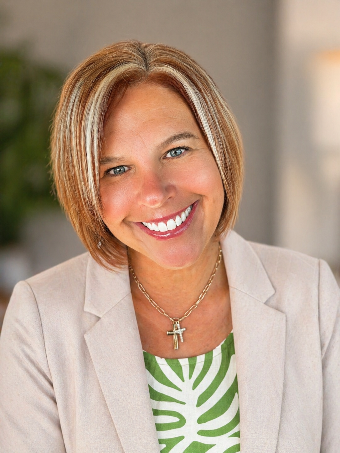 A smiling woman with short blonde hair and blue eyes, wearing a beige blazer, a green and white patterned top, and a necklace with cross pendants.