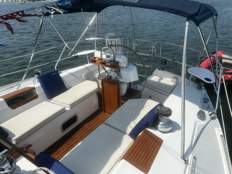The deck of a sailboat with seating, cushions, a small table, and sailing equipment covered by a blue canopy, floating on water.