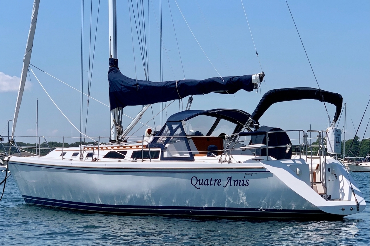 A white sailboat named Quatre Amis with a blue sail cover, moored on the water on a sunny day with a clear blue sky, multiple masts, and other boats in the background.