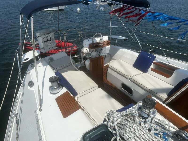 The image shows the deck of a sailboat with white cushioned seating, wood accents, and various nautical equipment. There are ropes, winches, a bimini top with colorful bunting, and a view of the water in the background.