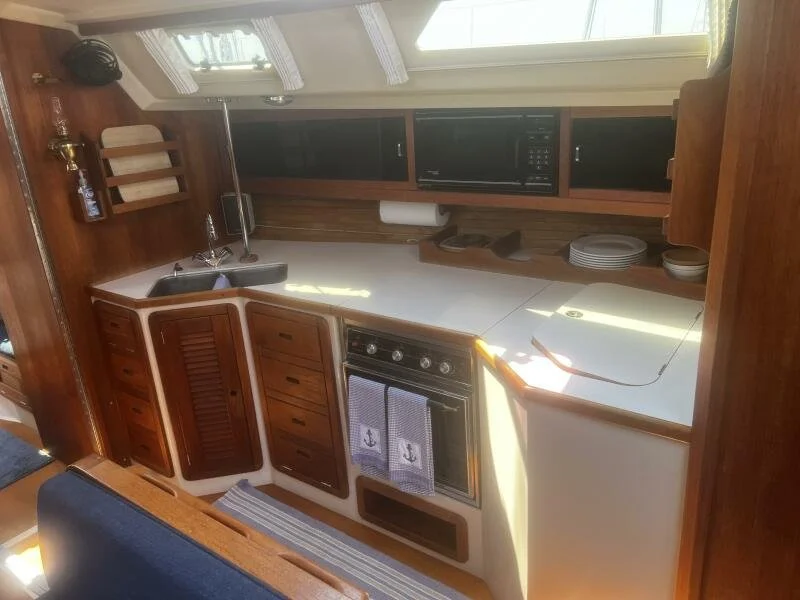 Compact kitchen with wooden cabinets, a stainless steel sink, stove, microwave, and various dishes, located in a boat or RV with skylight and window overhead.