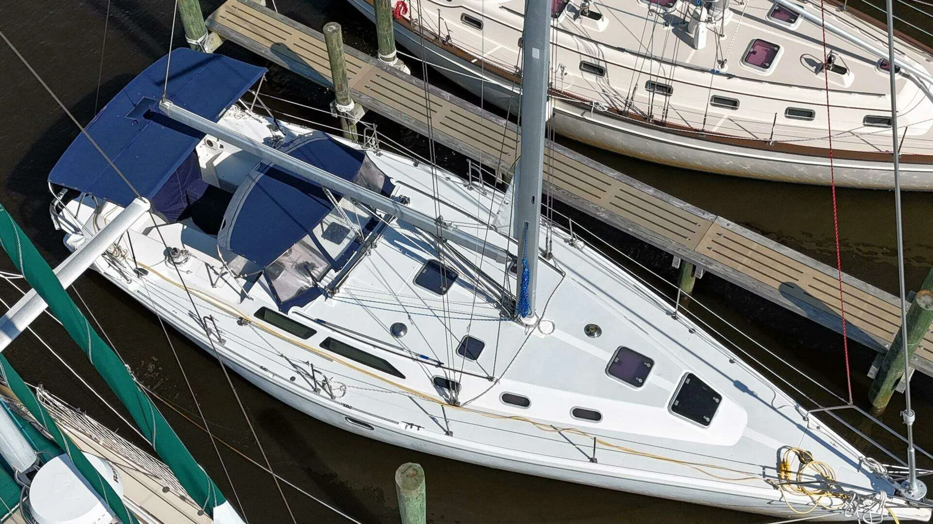A top-down view of a white sailboat docked at a marina, with a blue canopy over the cockpit, surrounded by other boats and a wooden pier