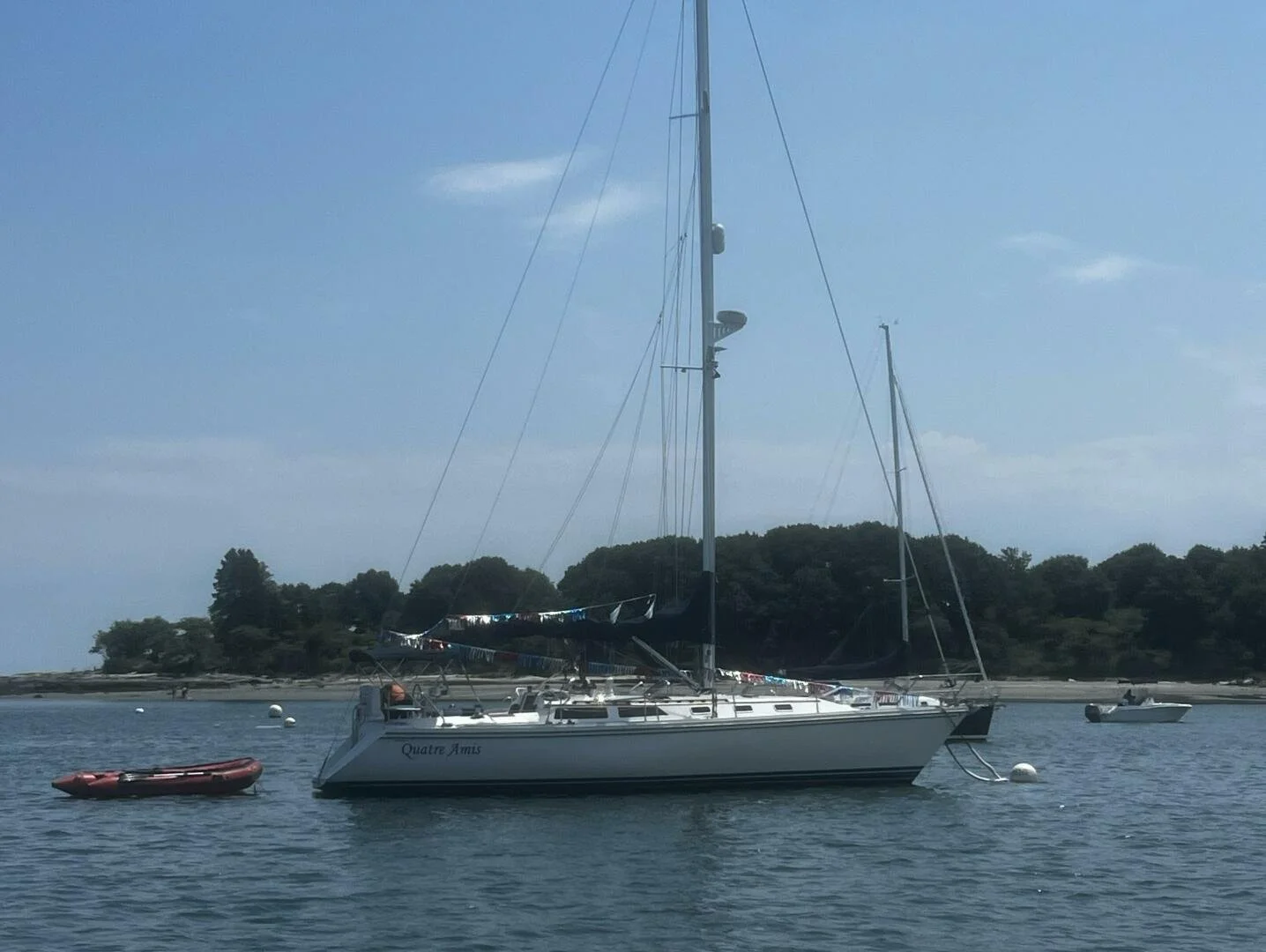 Sailboat named Quatre Amis anchored near a shoreline with trees, with a smaller boat, a kayak, and另外boats in the background.