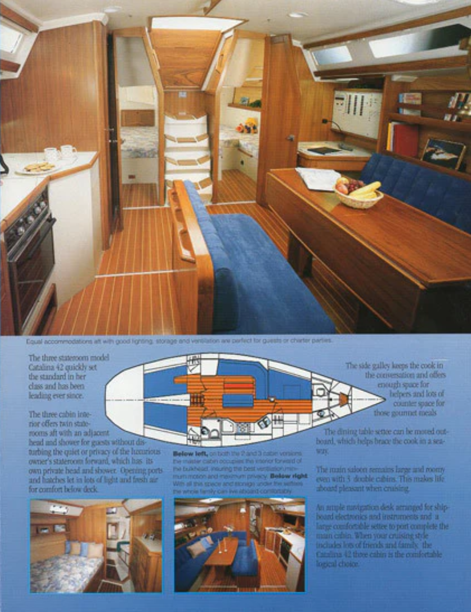 Interior of a boat's living area with wooden walls and blue upholstered seating around a dining table, a small kitchen to the side, and a sleeping area in the back with a bed and shelves. A fruit basket is on the table.