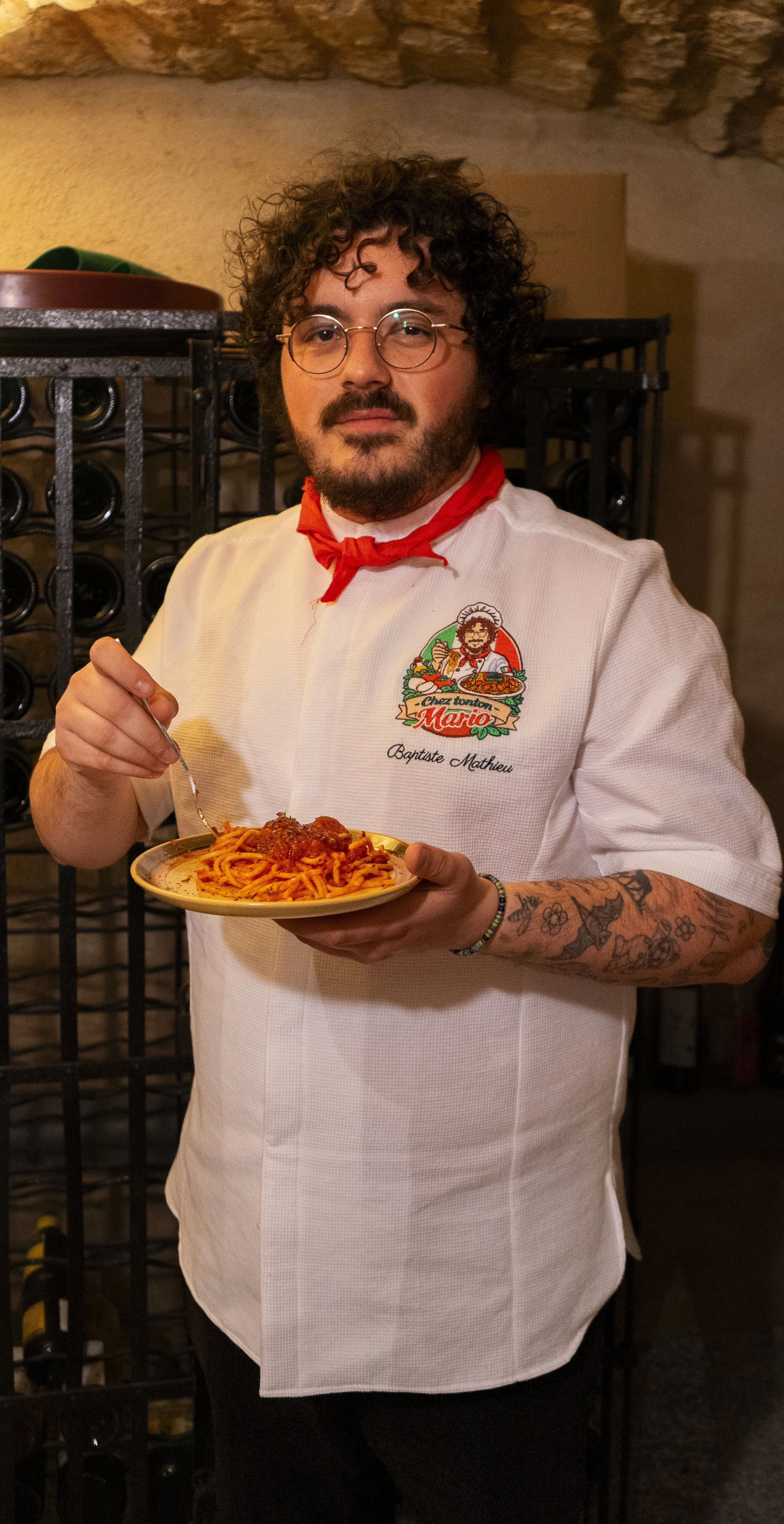 Un homme avec des tatouages portant une veste blanche avec un logo de restaurant et une barbe, tenant une assiette de spaghetti à la bolognaise, dans un restaurant.