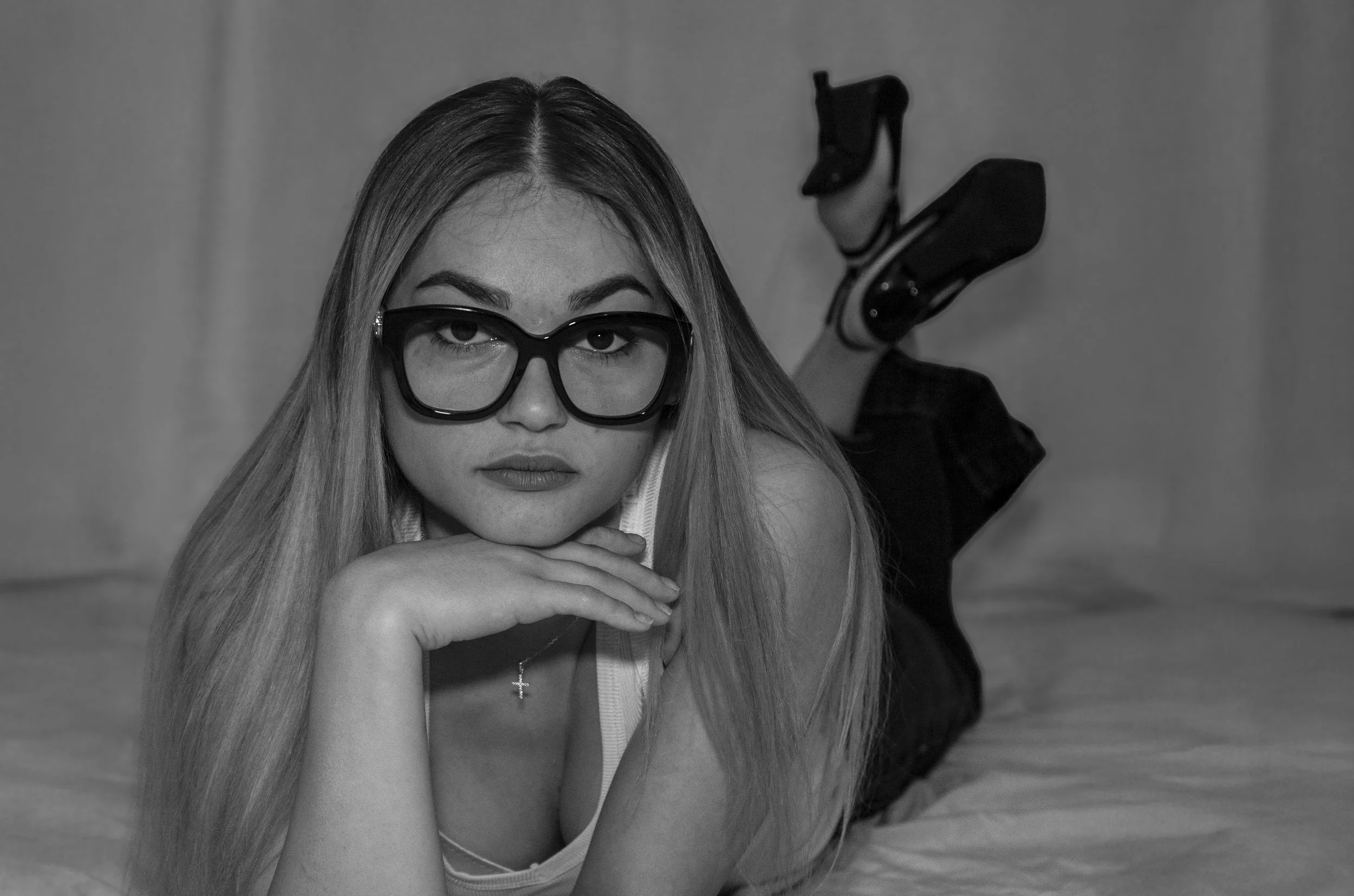 Une jeune femme aux longs cheveux blonds portant de grosses lunettes noires, posant avec son visage appuyé sur sa main. Elle porte un collier avec une croix et une chemise blanche à rayures. L'arrière-plan est neutre, et elle regarde directement l'objectif.