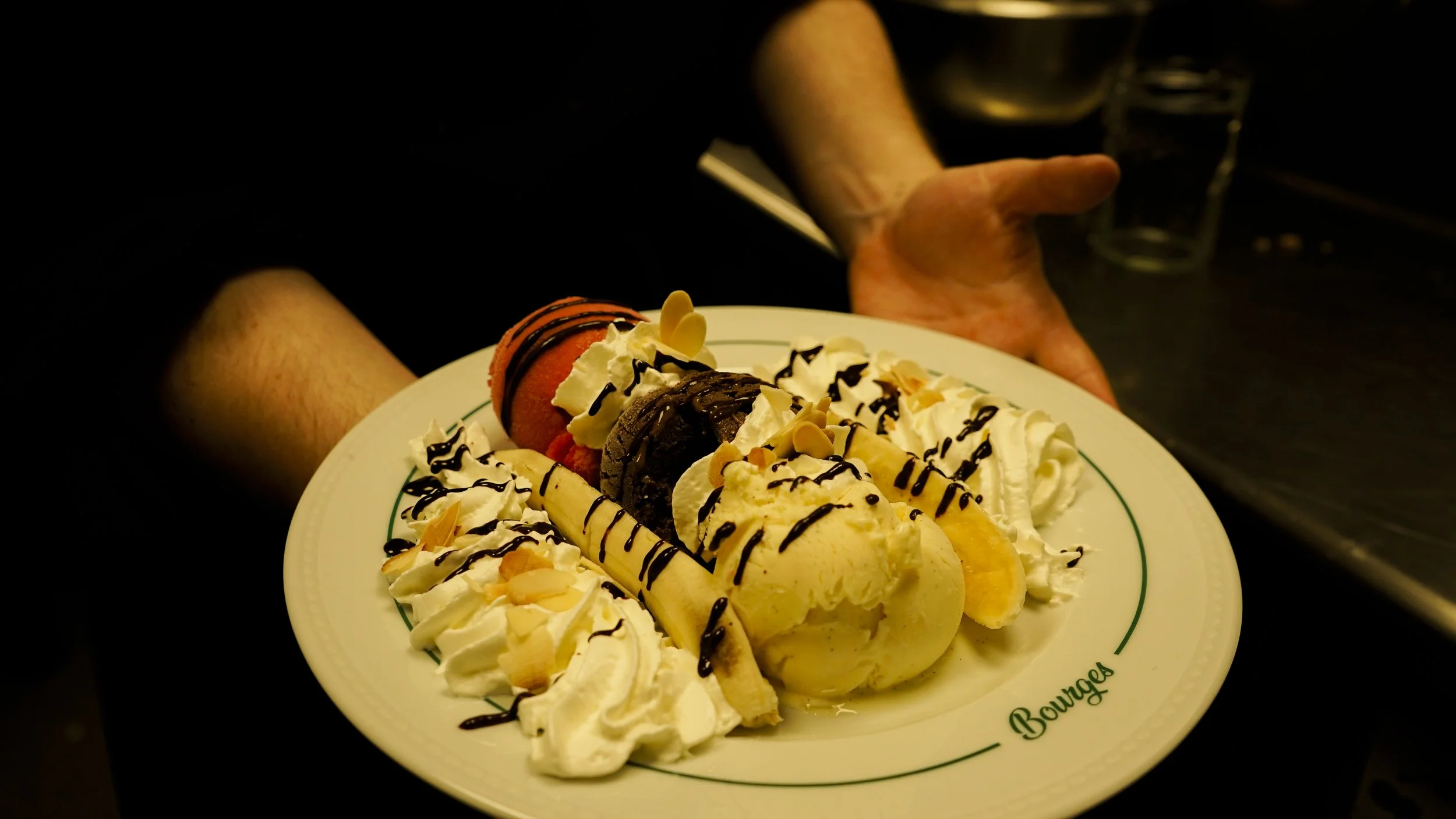 Assiette de glace à la vanille garnie de chantilly, chocolat fondu, amandes et fruits, tenue par une personne, fond sombre en arrière-plan.