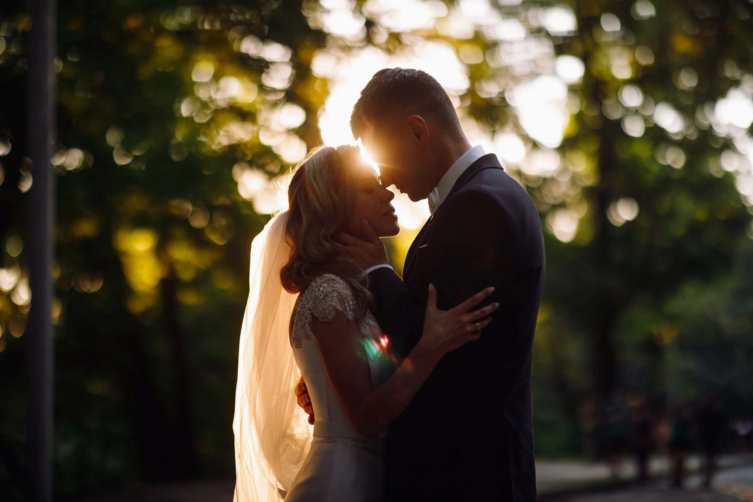 Un couple en mariage se tient très proche, face à face, dans une lumière de coucher de soleil avec un arrière-plan flou de feuillages, partageant un moment intime.