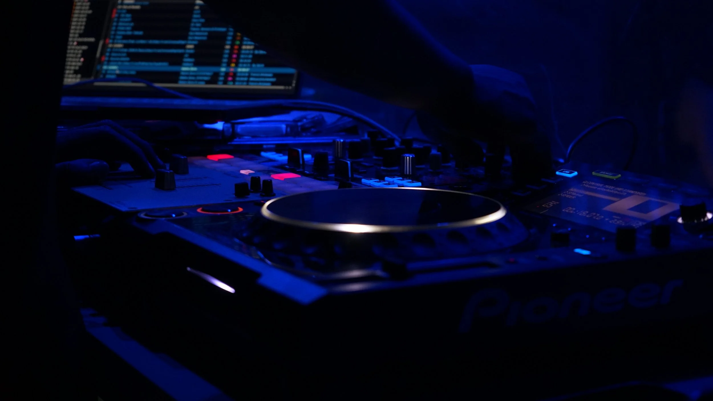 Plateforme de DJ avec console, contrôleurs, écran affichant une liste de chansons, éclairage bleu dans une ambiance sombre.