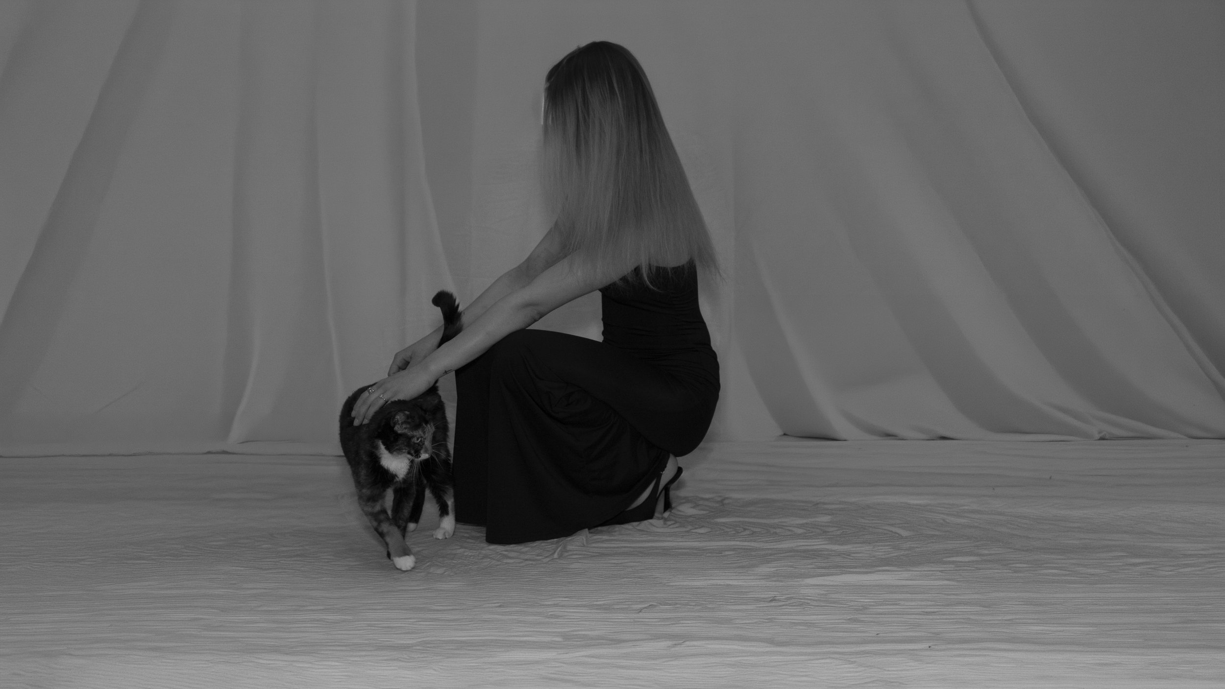 Une femme aux cheveux longs, habillée en noir, assise au sol en face d’un chat noir et blanc. Le décor est monochrome avec des drapés blancs en fond.