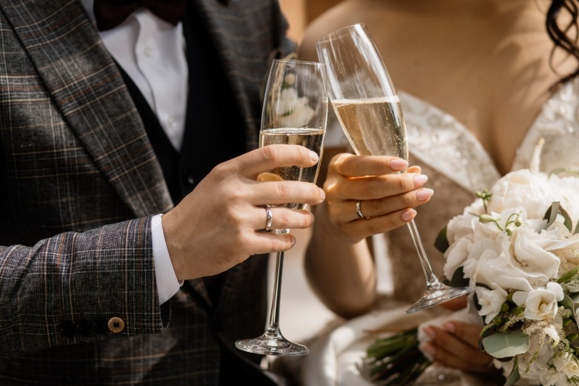 Un couple à un mariage, ils tiennent des flûtes de champagne toastant, la femme tient un bouquet de fleurs blanches.