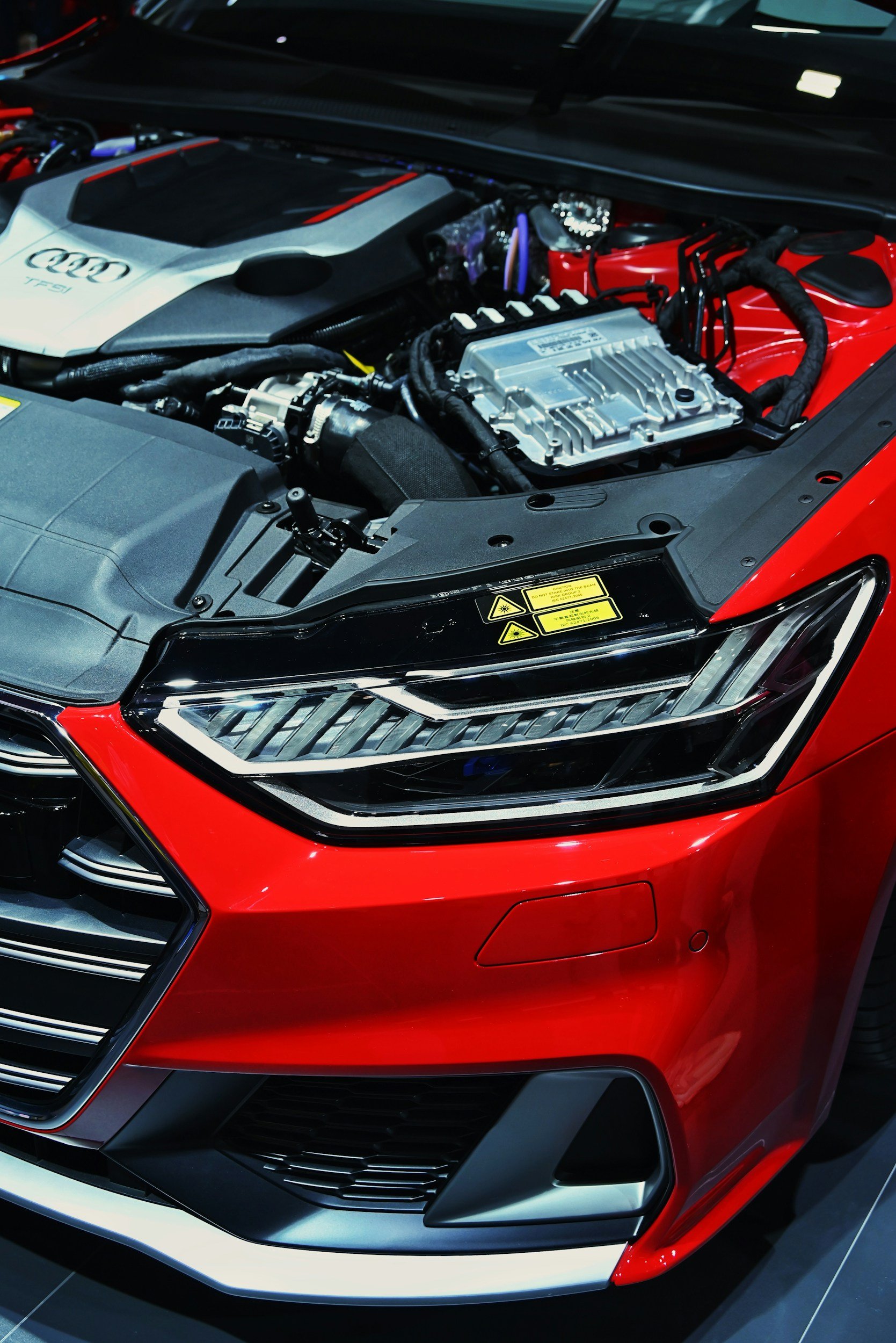 Close-up of a red and black modern car headlight and front grille with an opened hood revealing the engine compartment.