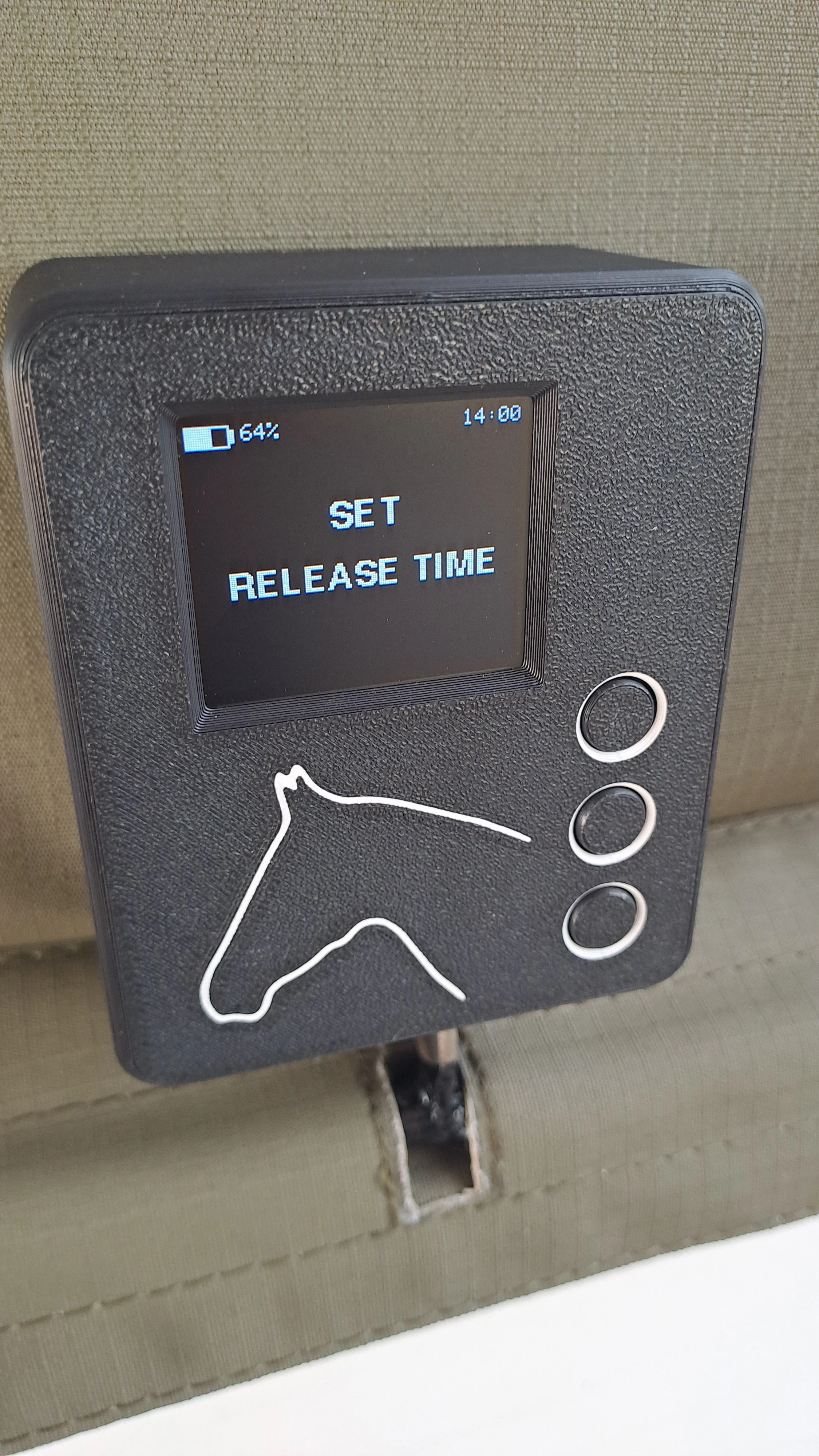 A digital device with a small screen displaying 'SET RELEASE TIME,' with a low battery icon and time 14:00, mounted on a textured surface. The device has a white horse outline and three round buttons.