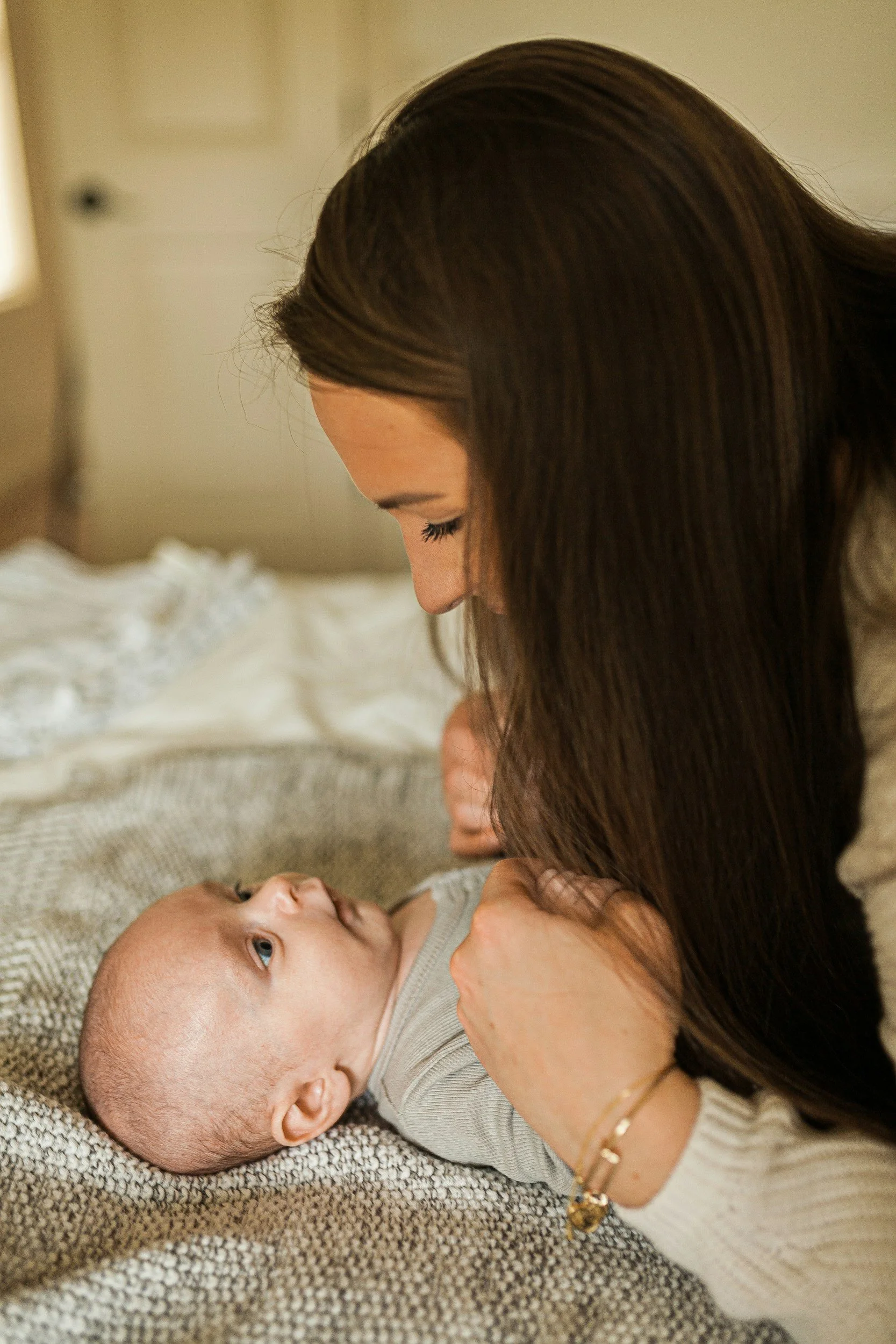 A woman with long brown hair lies on a bed, looking down at and gently holding a sleeping baby's face. The baby, with blue eyes and wearing a gray shirt, is lying on a textured blanket and gazing up at the woman.