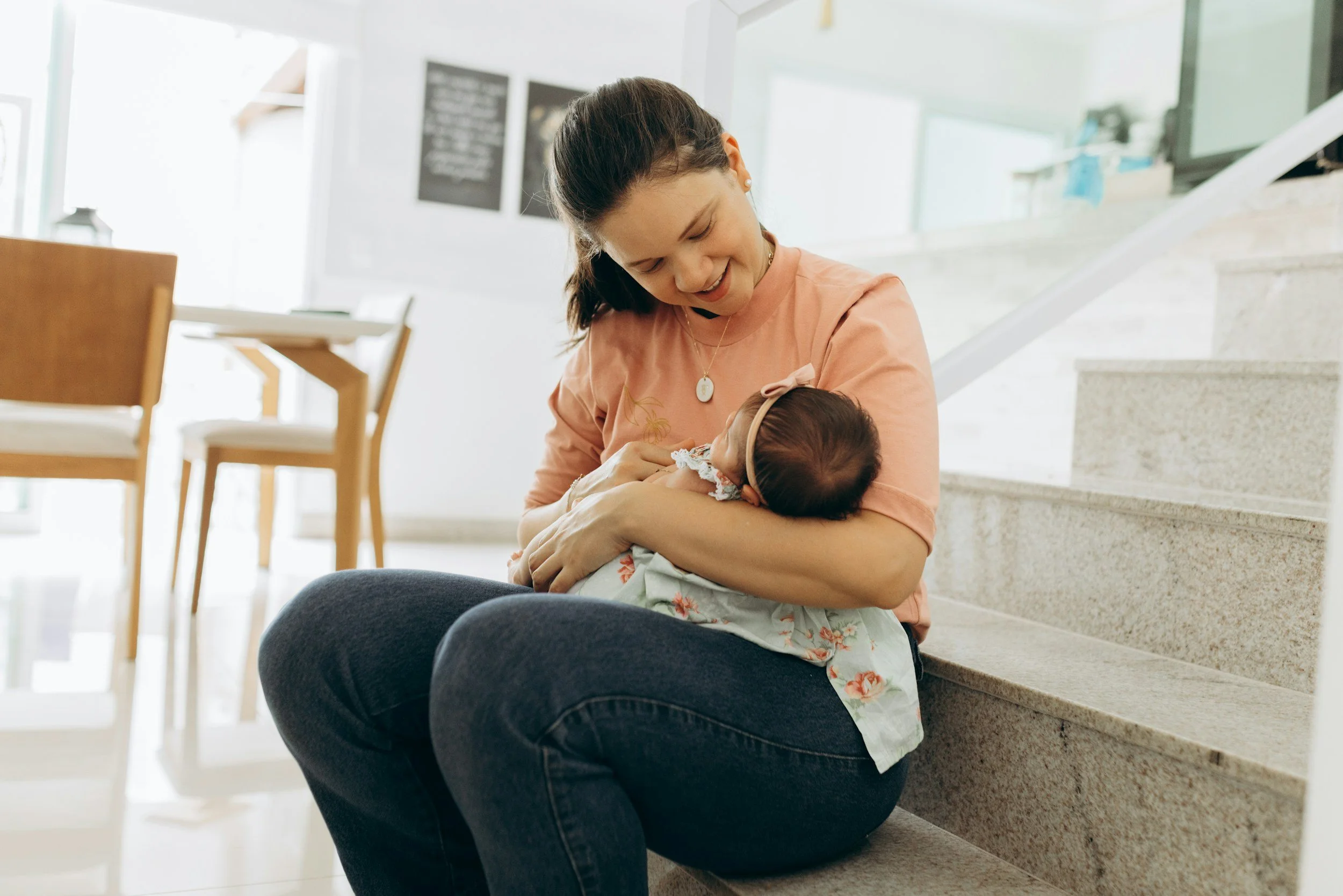 A woman holding a baby and sitting on a sofa, smiling at another woman who is sitting on the other side of the sofa with a notebook and pen, in a cozy living room with sunlight coming through the window. There is a framed sign on the wall that says 'You're doing a great job, mama.' on a side table, a plant, and a mug with a heart on it.