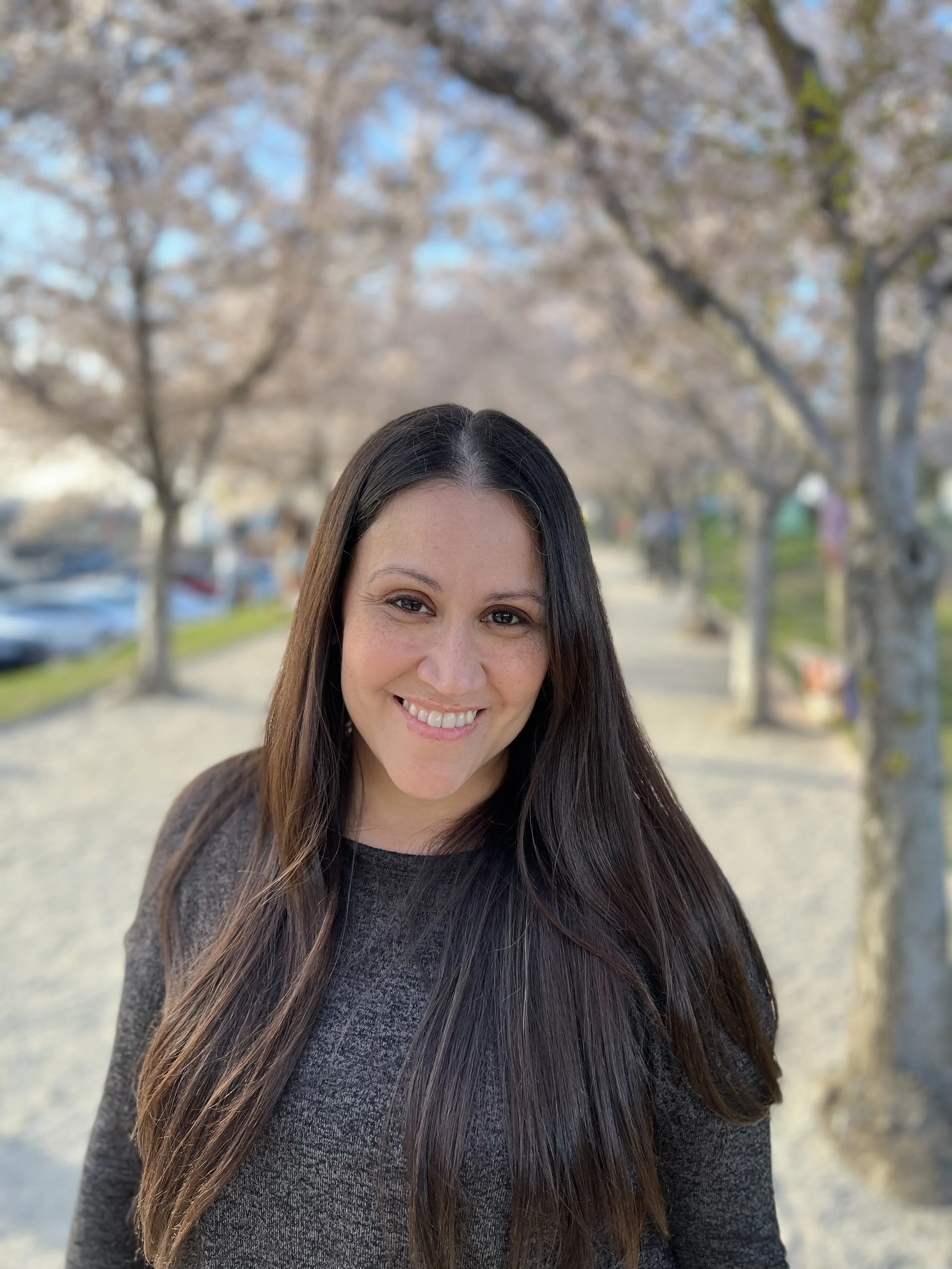 Close-up of a smiling woman with long dark hair, wearing a dark sweater, standing outdoors on a sunny day with trees and a sidewalk in the background.