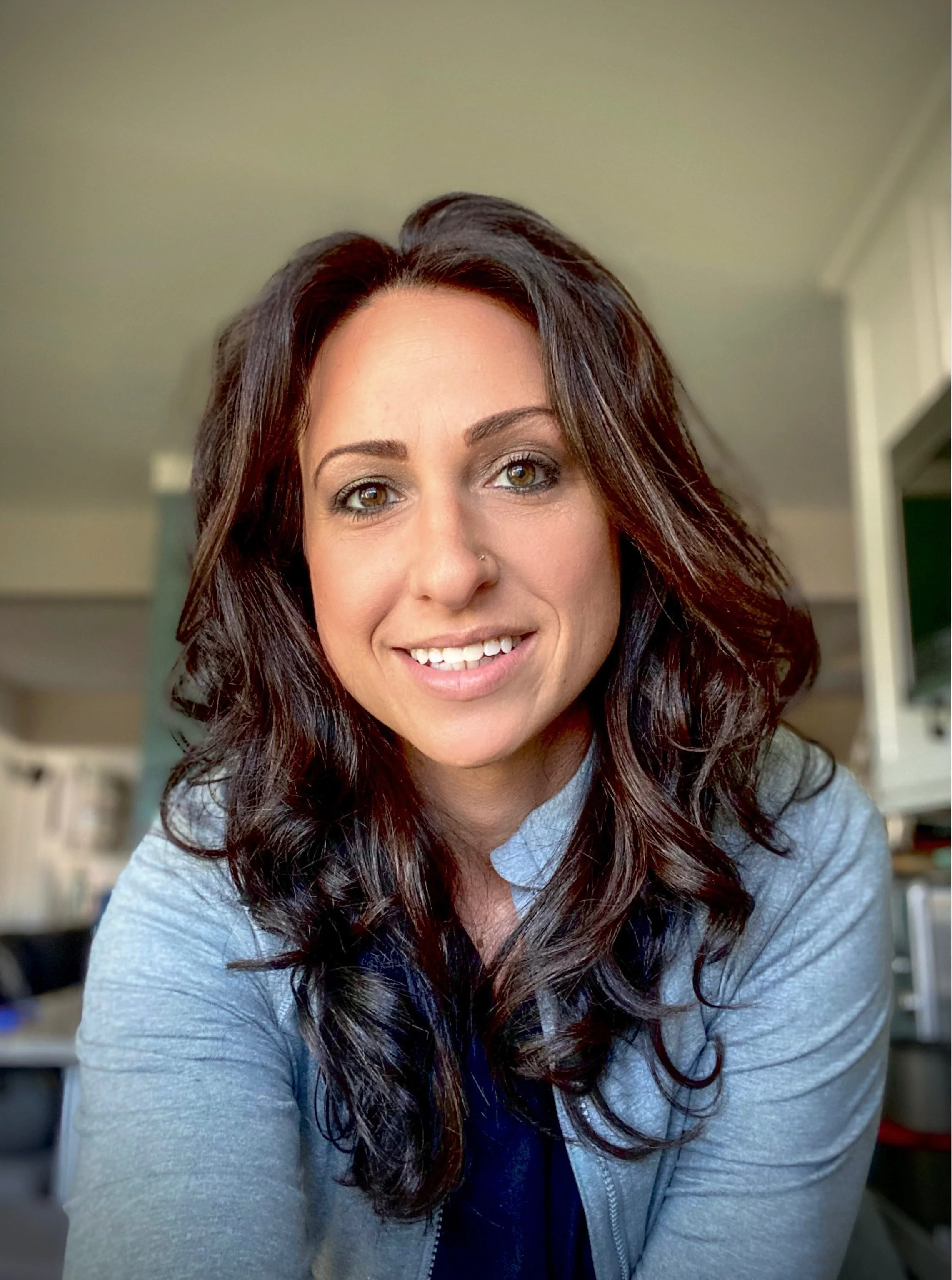 Close-up of a woman with long dark wavy hair, smiling, wearing a gray zip-up hoodie and a navy blue shirt underneath, indoors with a blurred background.