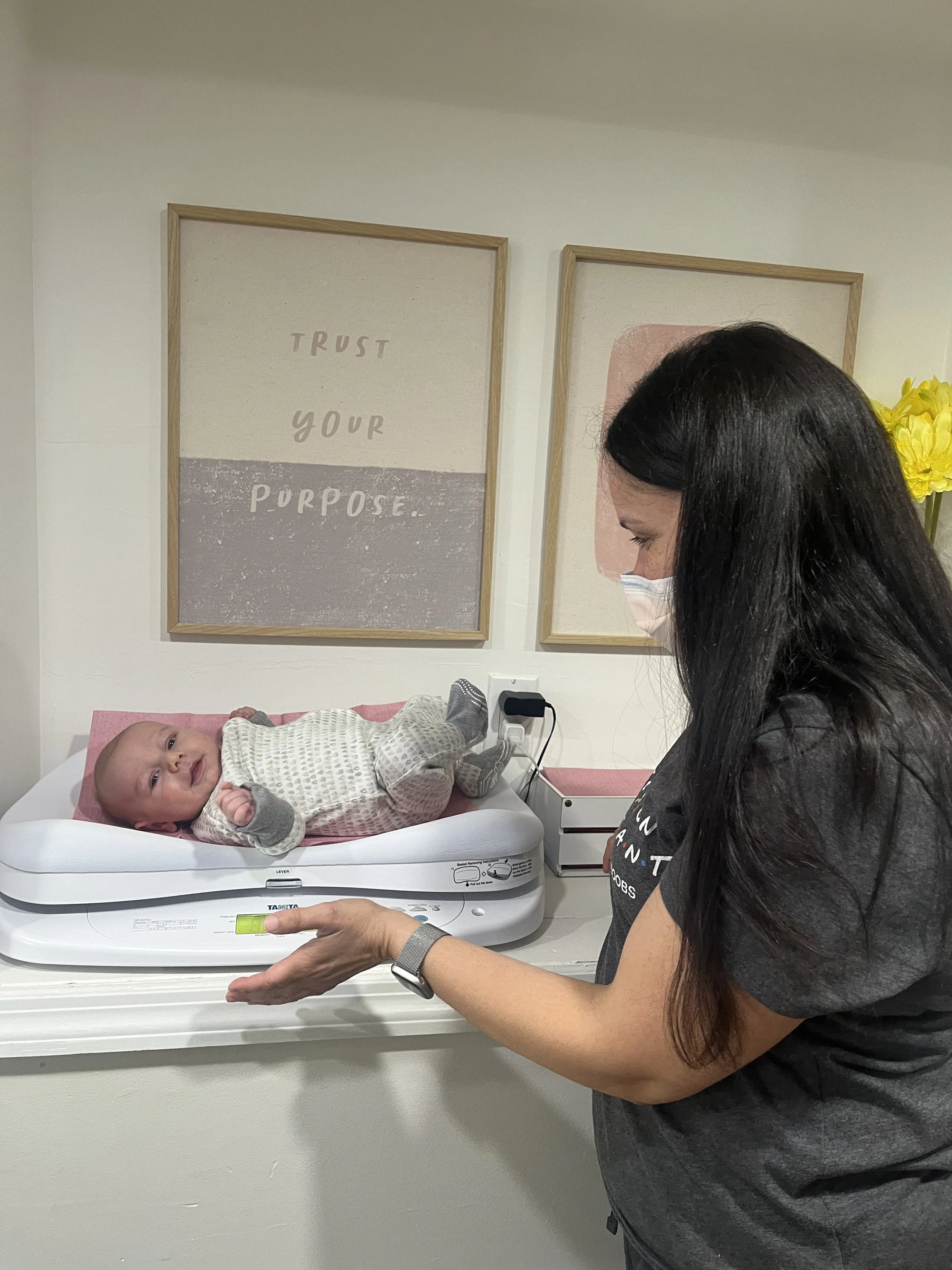 A woman is weighing a baby girl on a digital baby scale in a room with framed motivational posters on the wall that say 'TRUST YOUR PURPOSE'. The woman is wearing a face mask and a black shirt, and the baby girl is lying on her back, looking up at the camera.