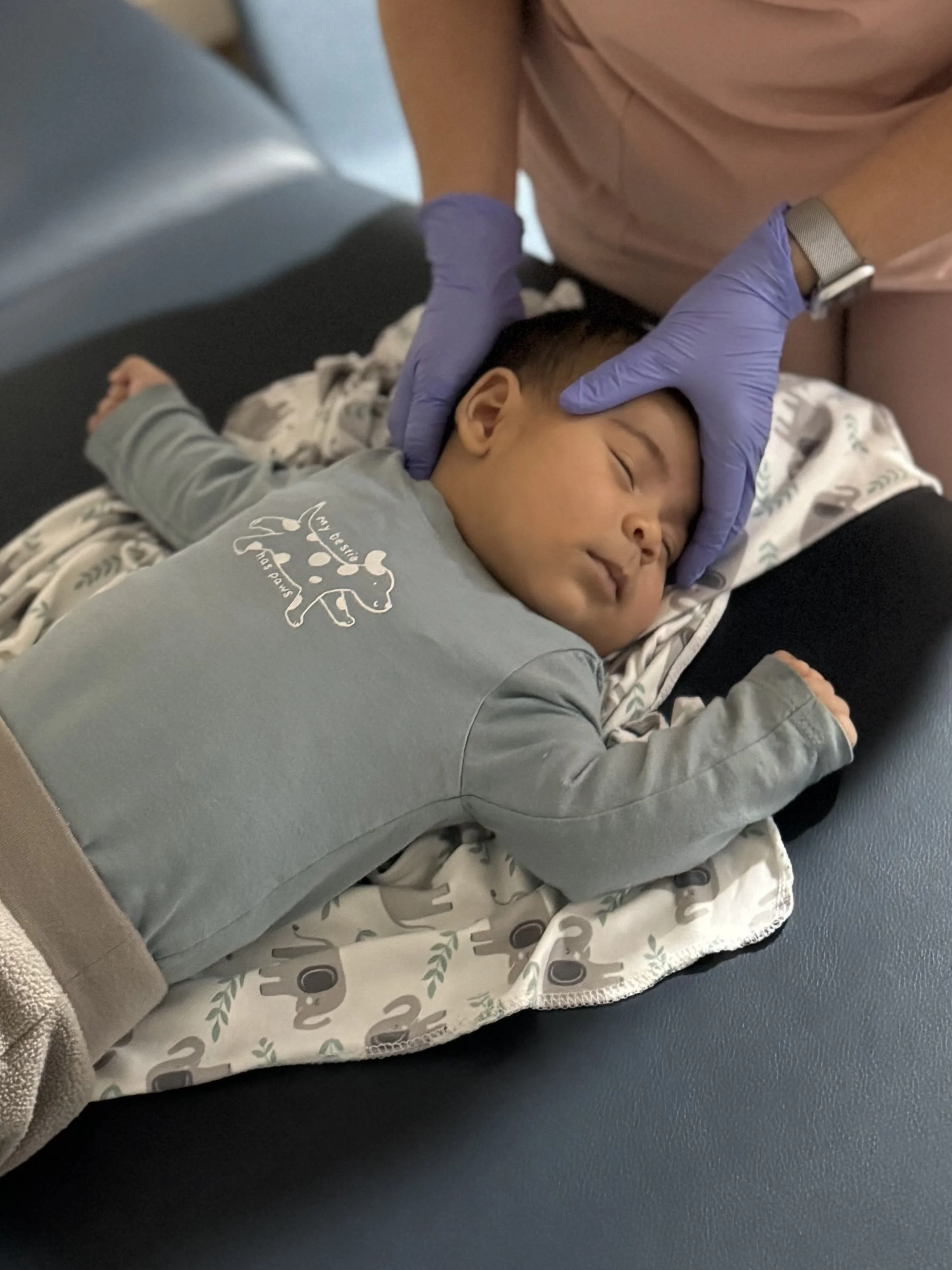 A young boy is lying on a hospital bed with medical gloves on the hands of a healthcare worker, who is checking his head. The boy appears relaxed and is resting peacefully.
