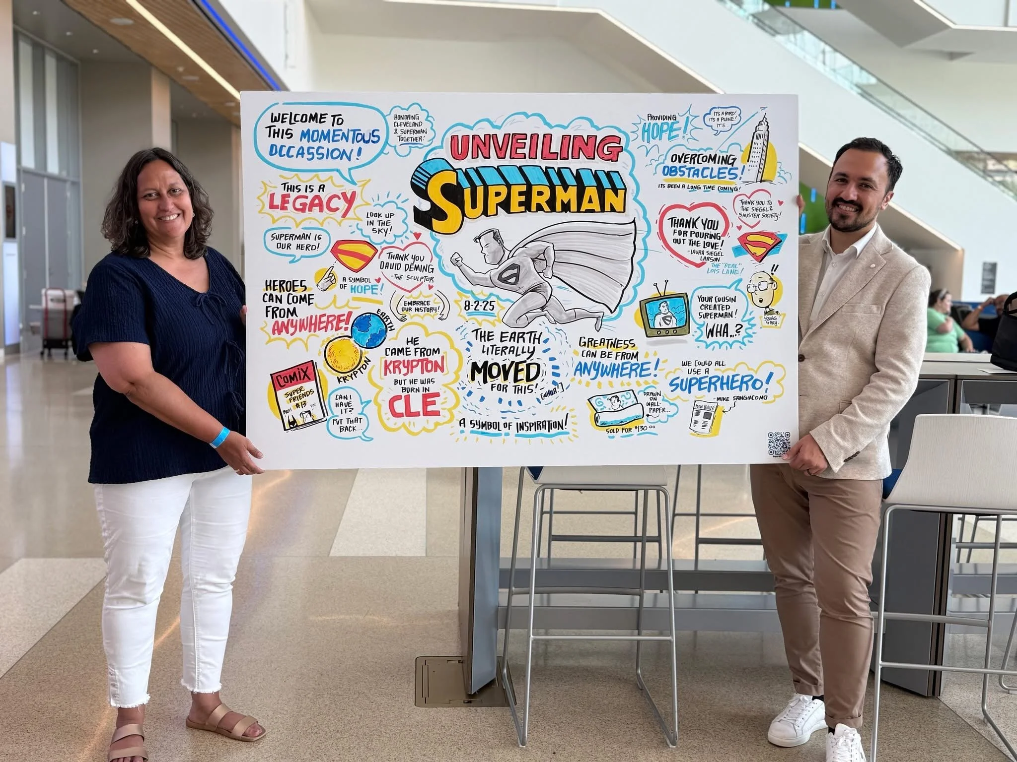 Two people, a woman on the left and a man on the right, are holding a large colorful poster about Superman. They are in a modern indoor space with glass walls and a high ceiling. The woman has shoulder-length brown hair, light skin, and is wearing a navy blue top with white pants. The man has dark hair, a beard, light skin, and is wearing a beige blazer with a white shirt and beige pants. The poster features drawings and text related to Superman, including quotes, symbols, and references to his origin and heroism.