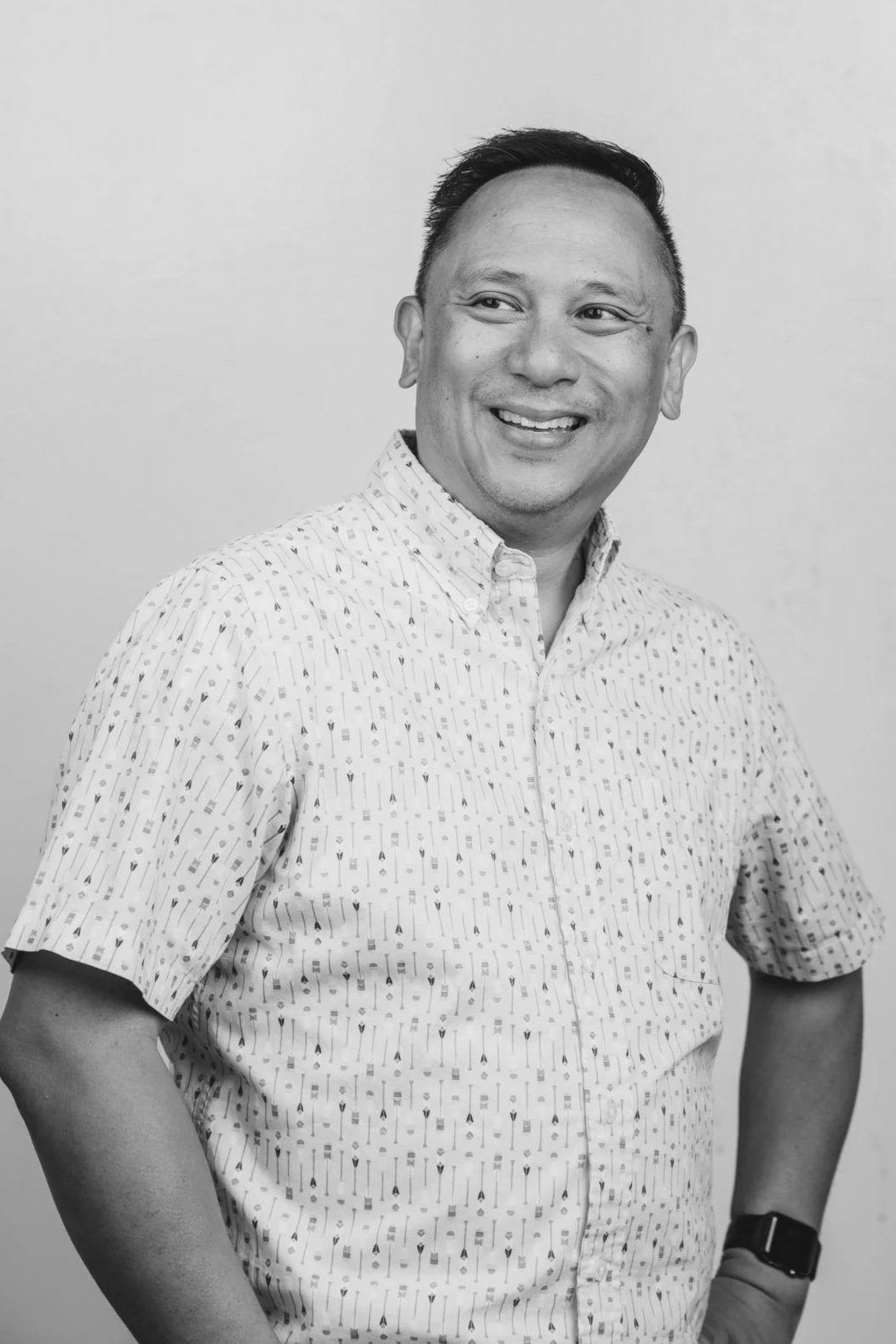 Black and white portrait of a man smiling, wearing a patterned short-sleeve button-up shirt and a watch, standing against a plain background.