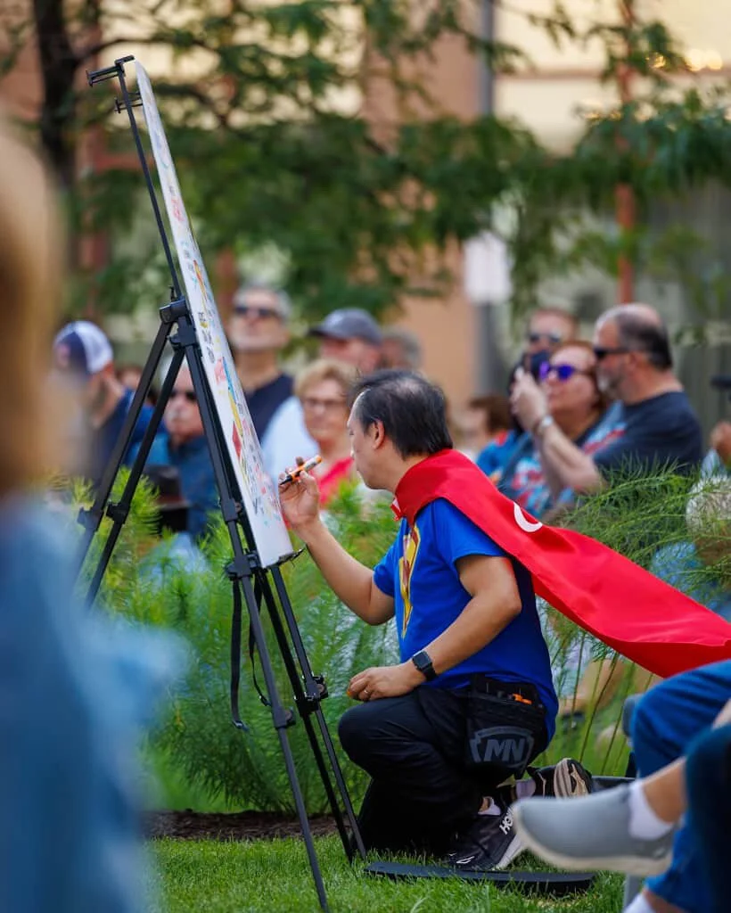 A man dressed as a superhero, wearing a red cape and a blue shirt, is painting a large outdoor mural or poster on an easel during a public event. The scene is set outdoors with a crowd of onlookers and greenery in the background.