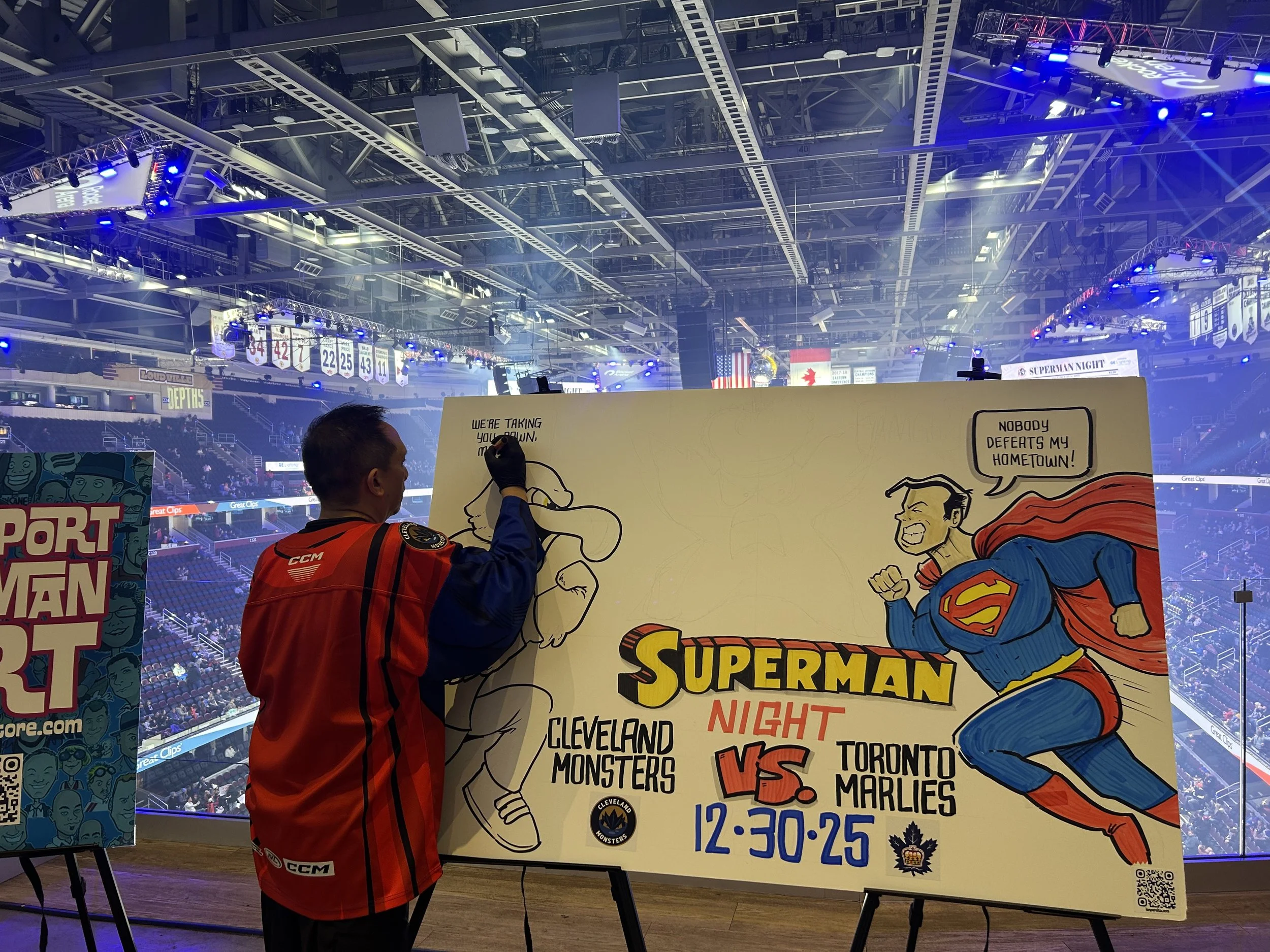 A person in a red hockey jersey drawing a cartoon of Superman on a large poster for a hockey game between the Cleveland Monsters and the Toronto Marlies at 12:30 PM. The background shows an indoor sports arena with banners, lighting, and spectators.