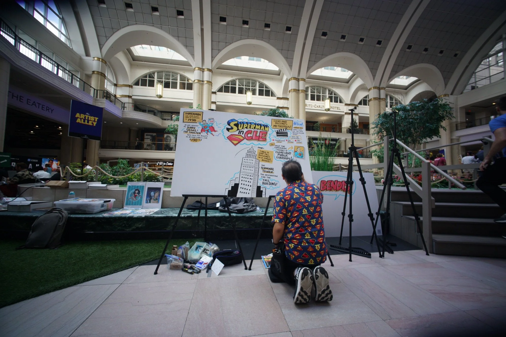 Person kneeling on the floor working on a large illustrated poster about Superman in Cleveland at an indoor shopping mall, with supplies and artwork on the table nearby, and a large open atrium with multiple floors and seating areas.