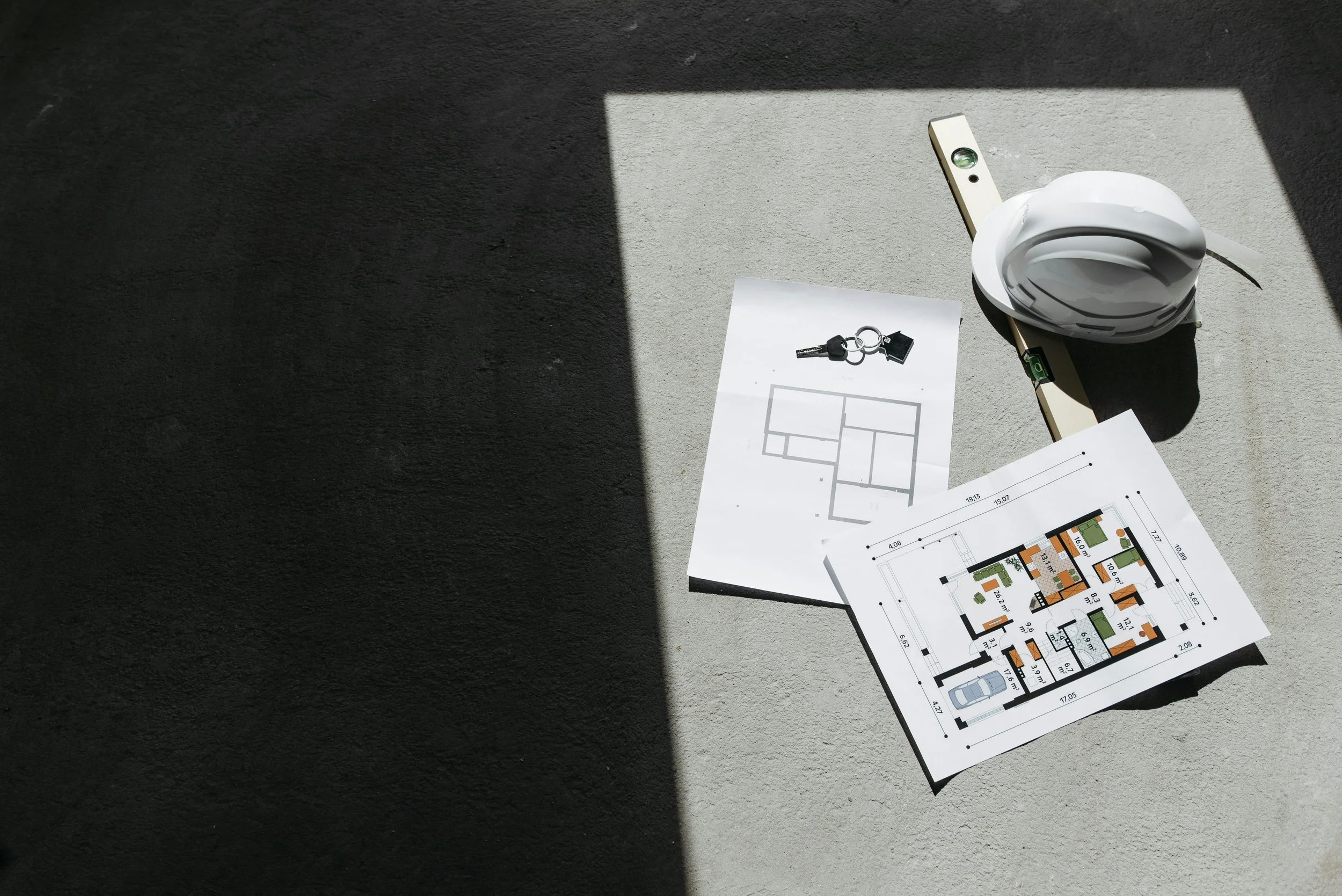Architectural blueprints and a white hard hat on a concrete surface, partially illuminated by sunlight.