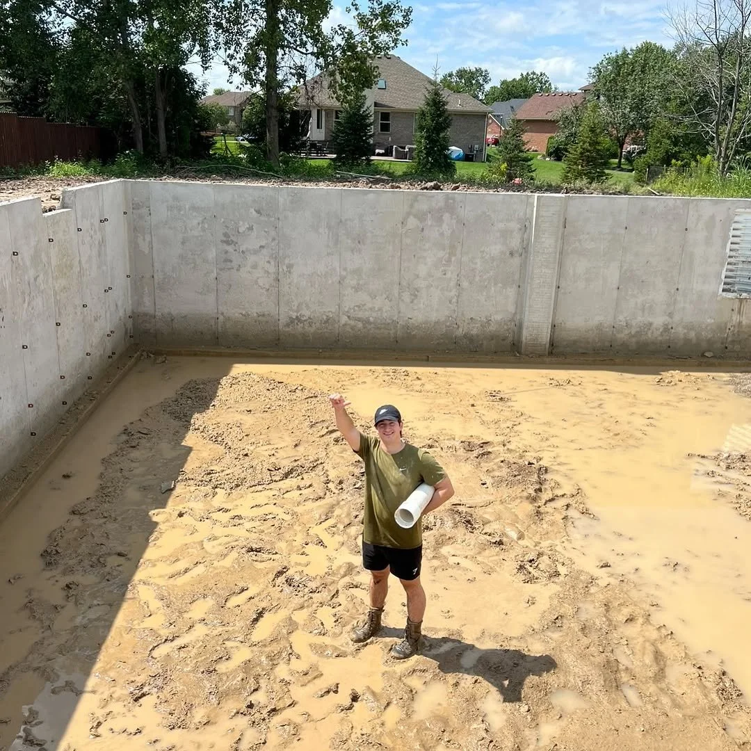 A person standing inside a concrete foundation with muddy ground, construction site, holding a rolled-up blueprint and raising one fist, smiling.
