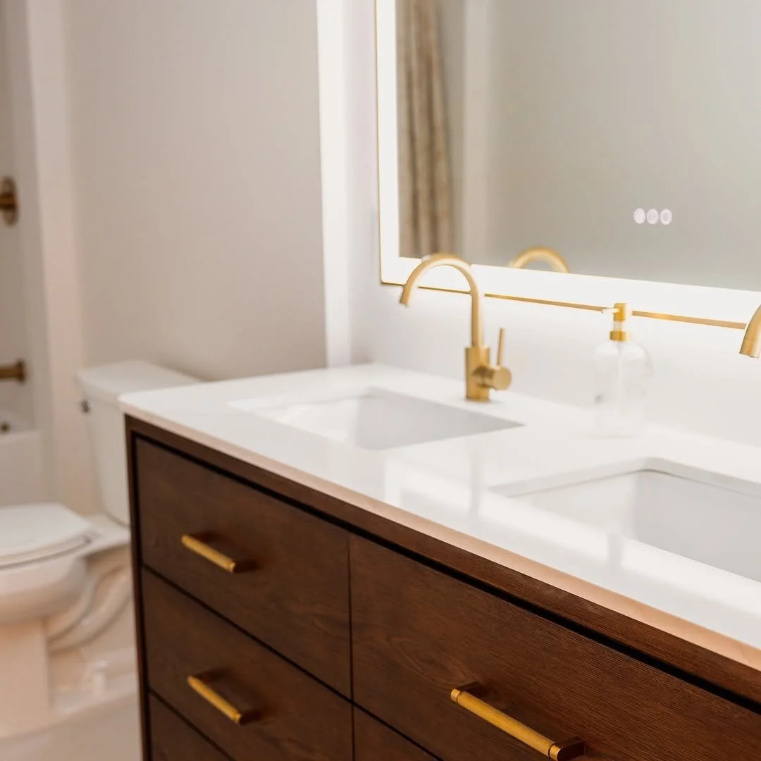 Bathroom with a white countertop, dual sinks with gold fixtures, a large mirror, a brown wooden vanity with gold handles, a clear soap dispenser, and a white toilet in the background.