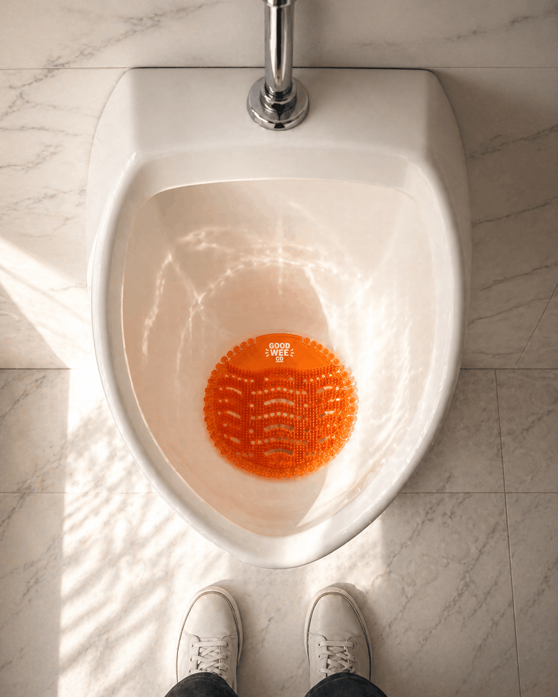 A person standing at a restroom urinal with feet visible, the urinal has an orange bright textured pad labeled "Good Wee Co" at the bottom.