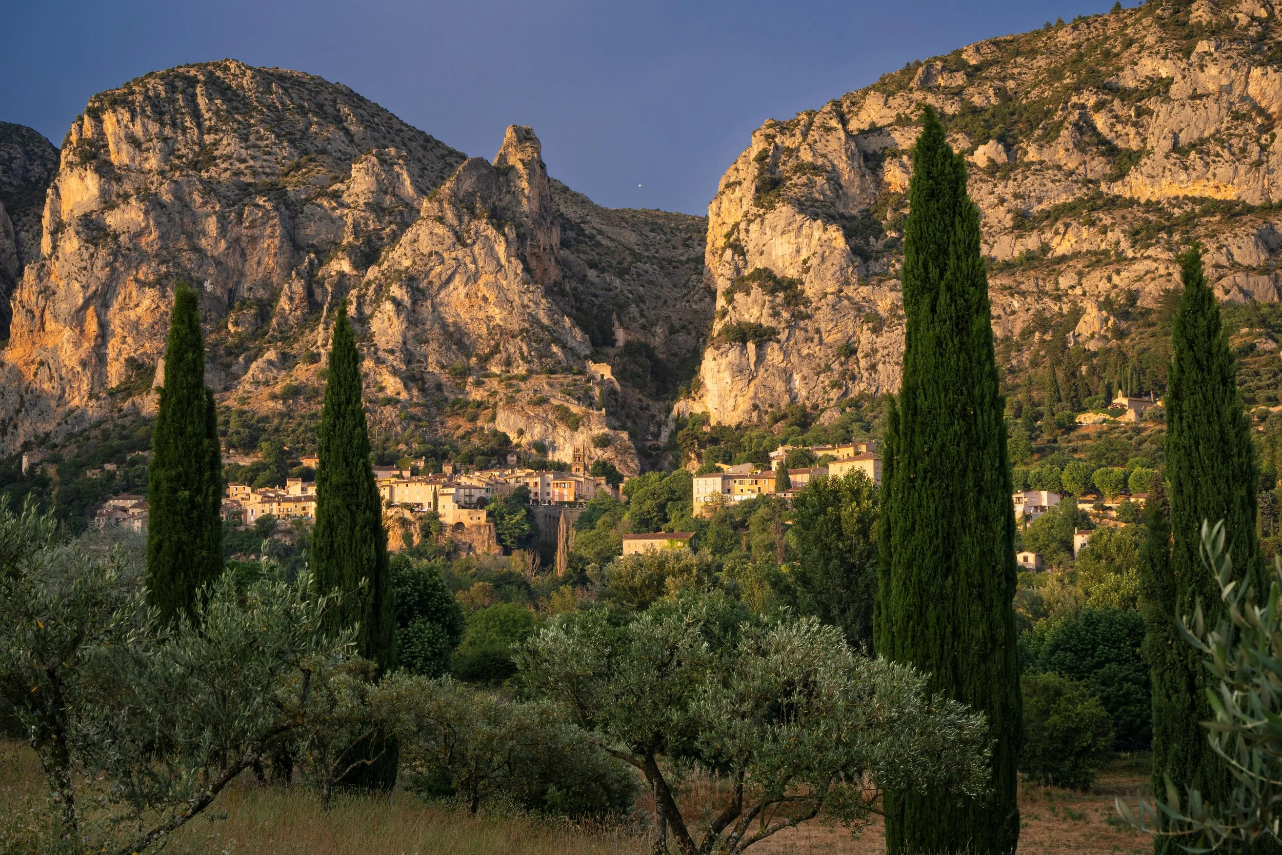 Paysage de montagnes rocheuses avec un village en contrebas et des arbres verts au premier plan.